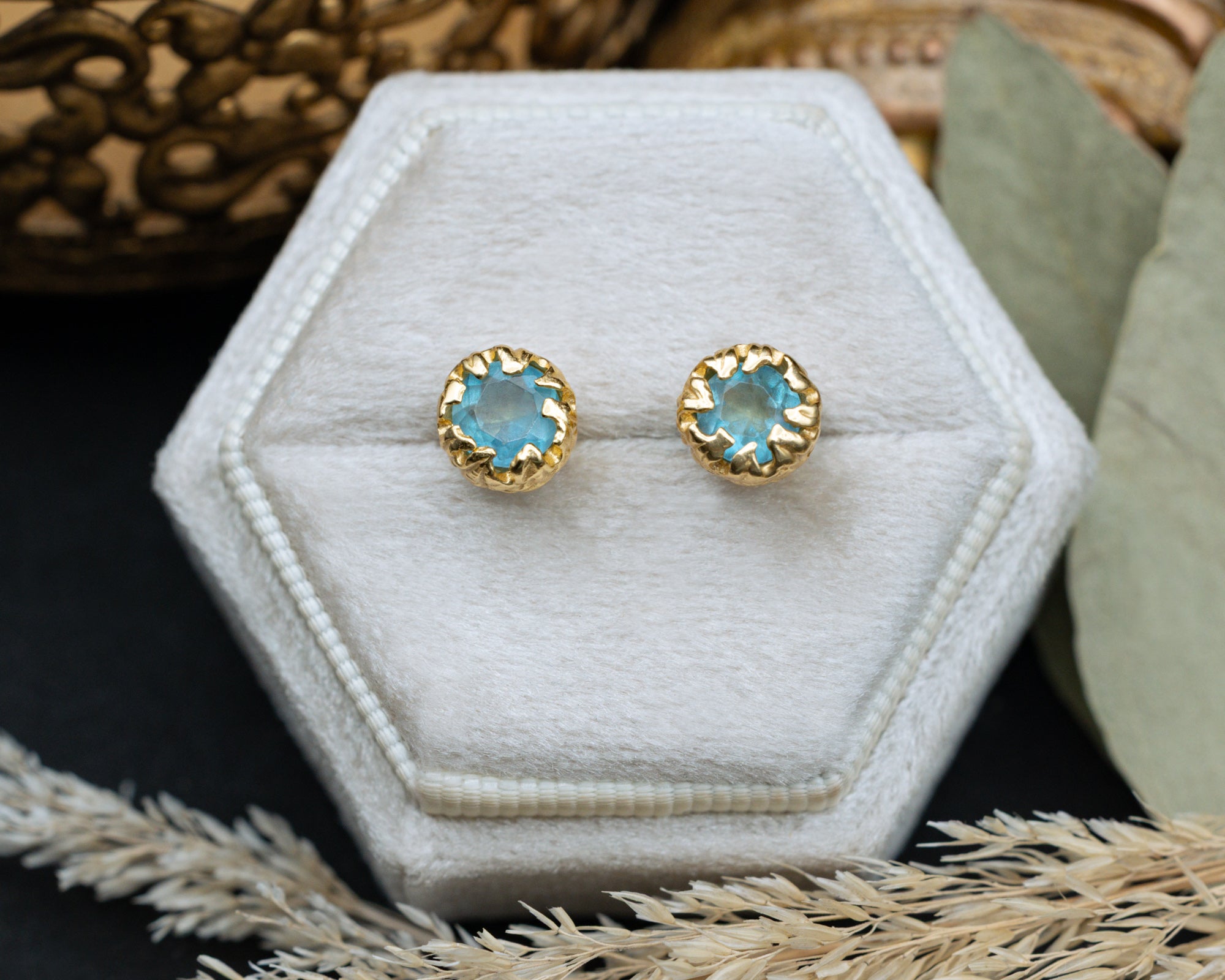 Gold earrings with blue aquamarine on a hexagonal white cushion, surrounded by natural elements.