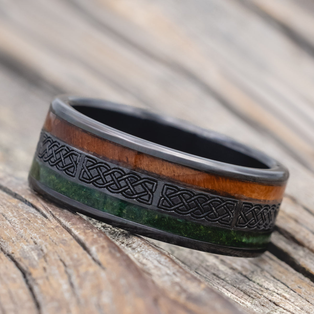 a black zirconium wedding ring featuring redwood and moss inlays with celtic engraving on a wooden surface