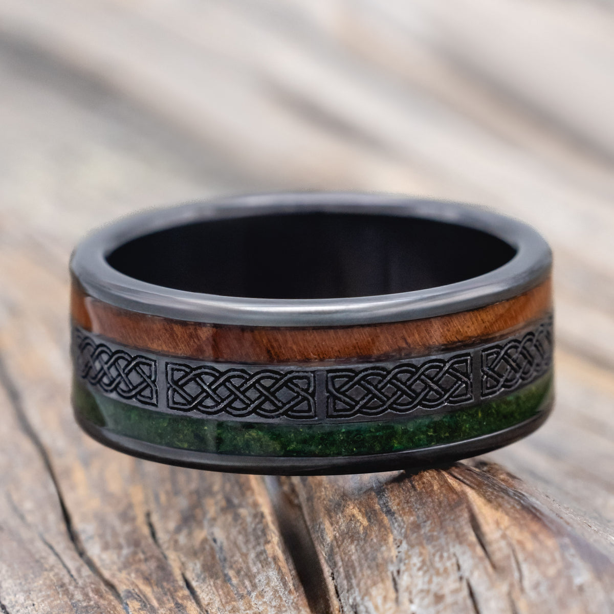 a black zirconium wedding ring featuring redwood and moss inlays with celtic engraving on a wooden surface