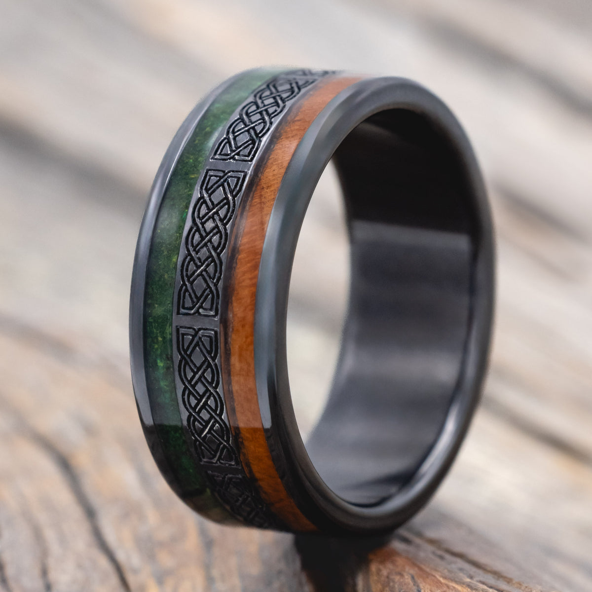 a black zirconium wedding ring featuring redwood and moss inlays with celtic engraving on a wooden surface
