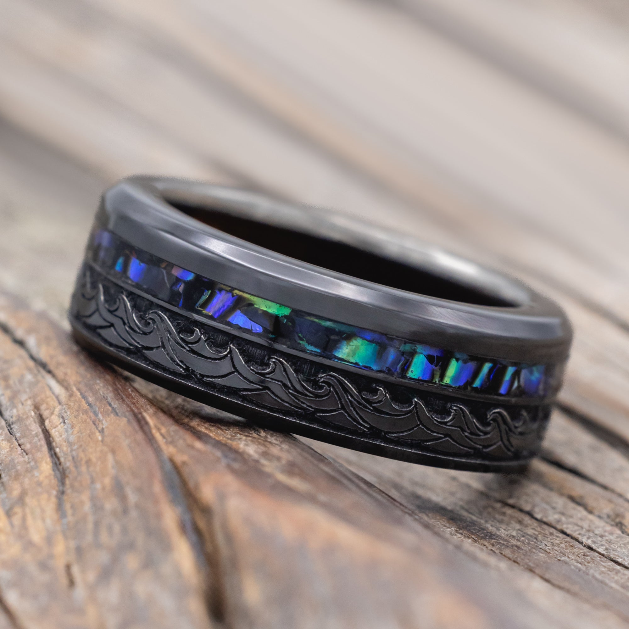 Black ring with paua shell, koa wood and wave engravings on a wooden surface