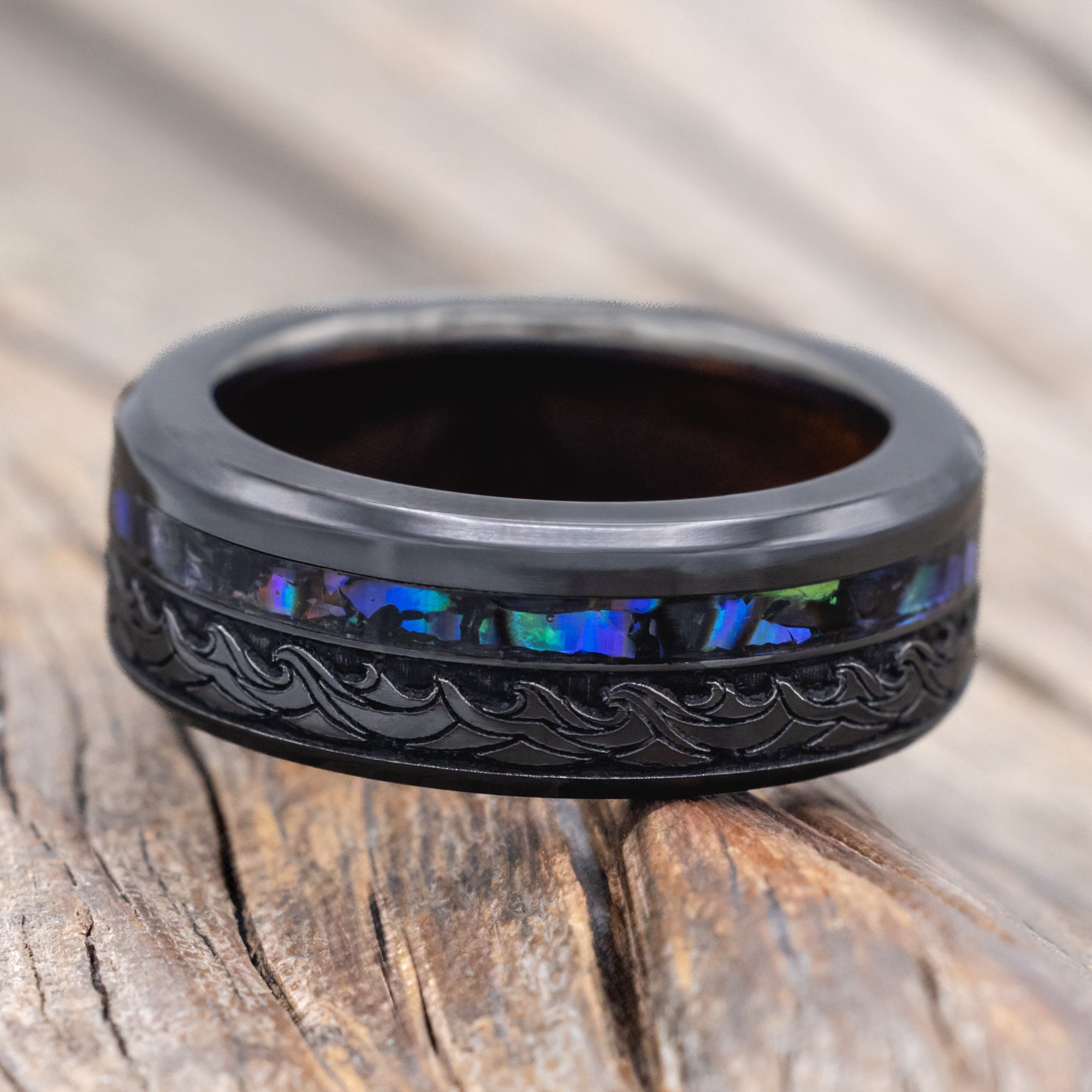 Black ring with paua shell, koa wood and wave engravings on a wooden surface