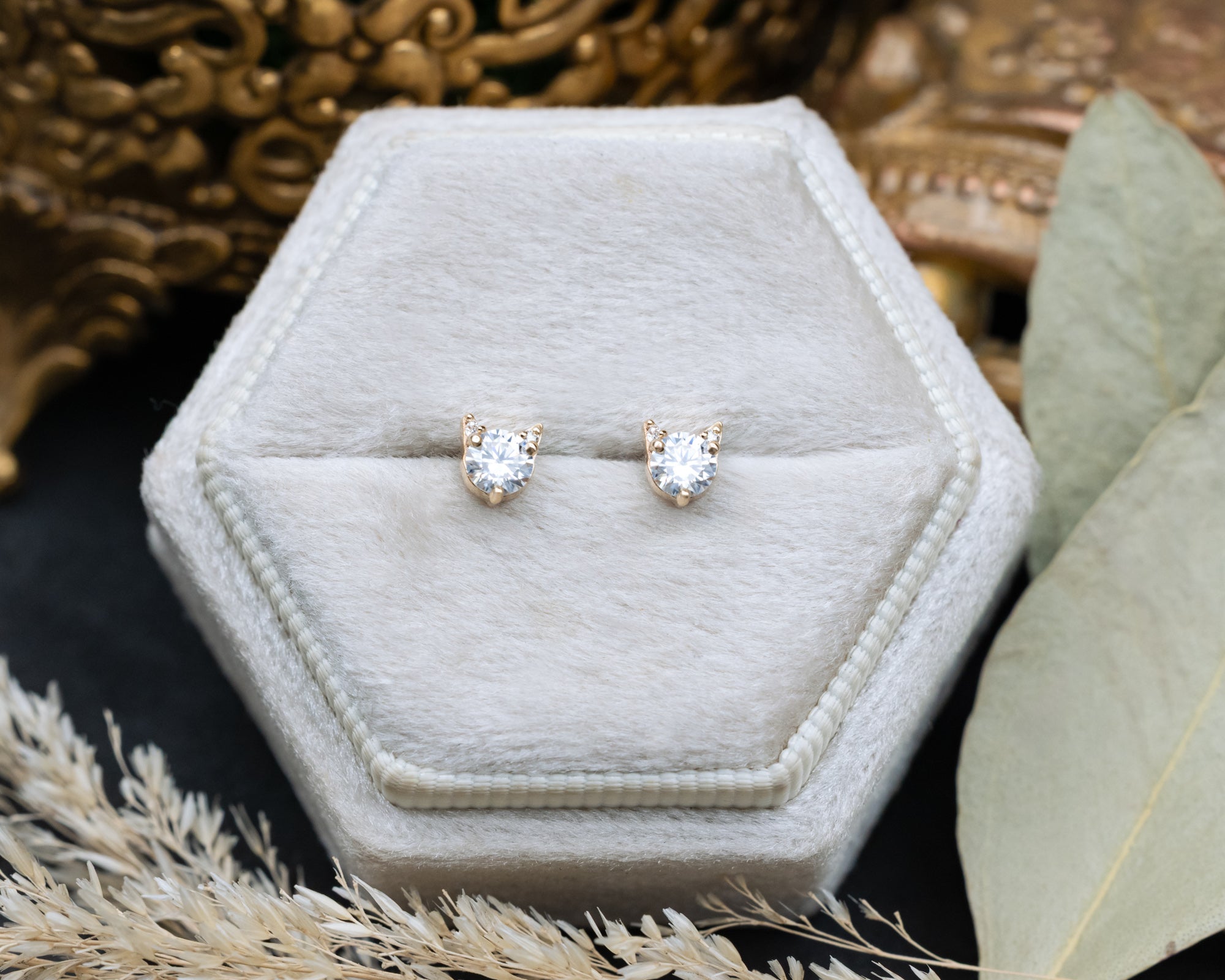 Pair of moissanite cat-shaped stud earrings on a white cushion against a decorative background