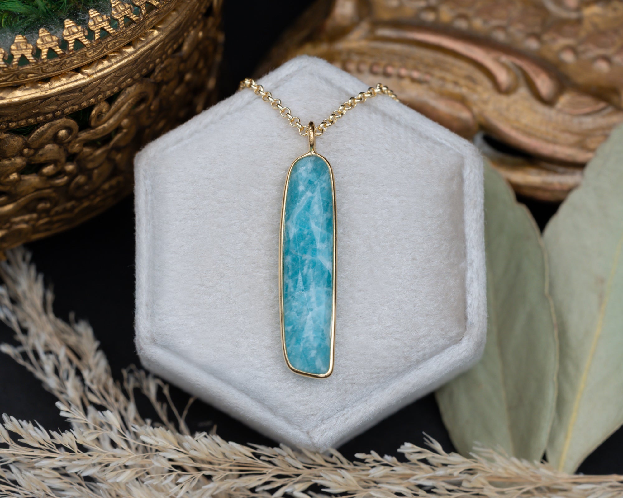 An amazonite pendant with a gold chain on a grey cushion with decorative elements in the background