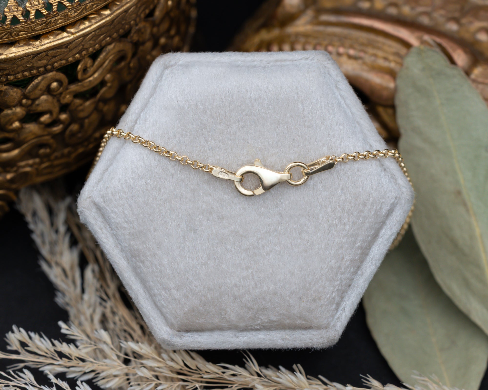 A gold chain with a lobster clasp, displayed on a gray velvet jewelry box with decorative dried grass and ornate gold-toned objects in the background.