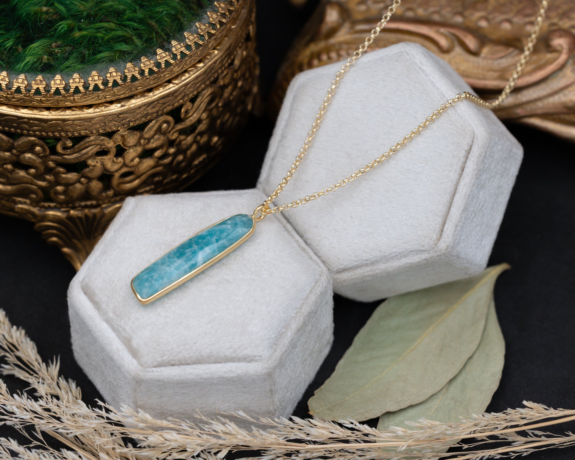 An amazonite pendant with a gold chain on a grey cushion with decorative elements in the background