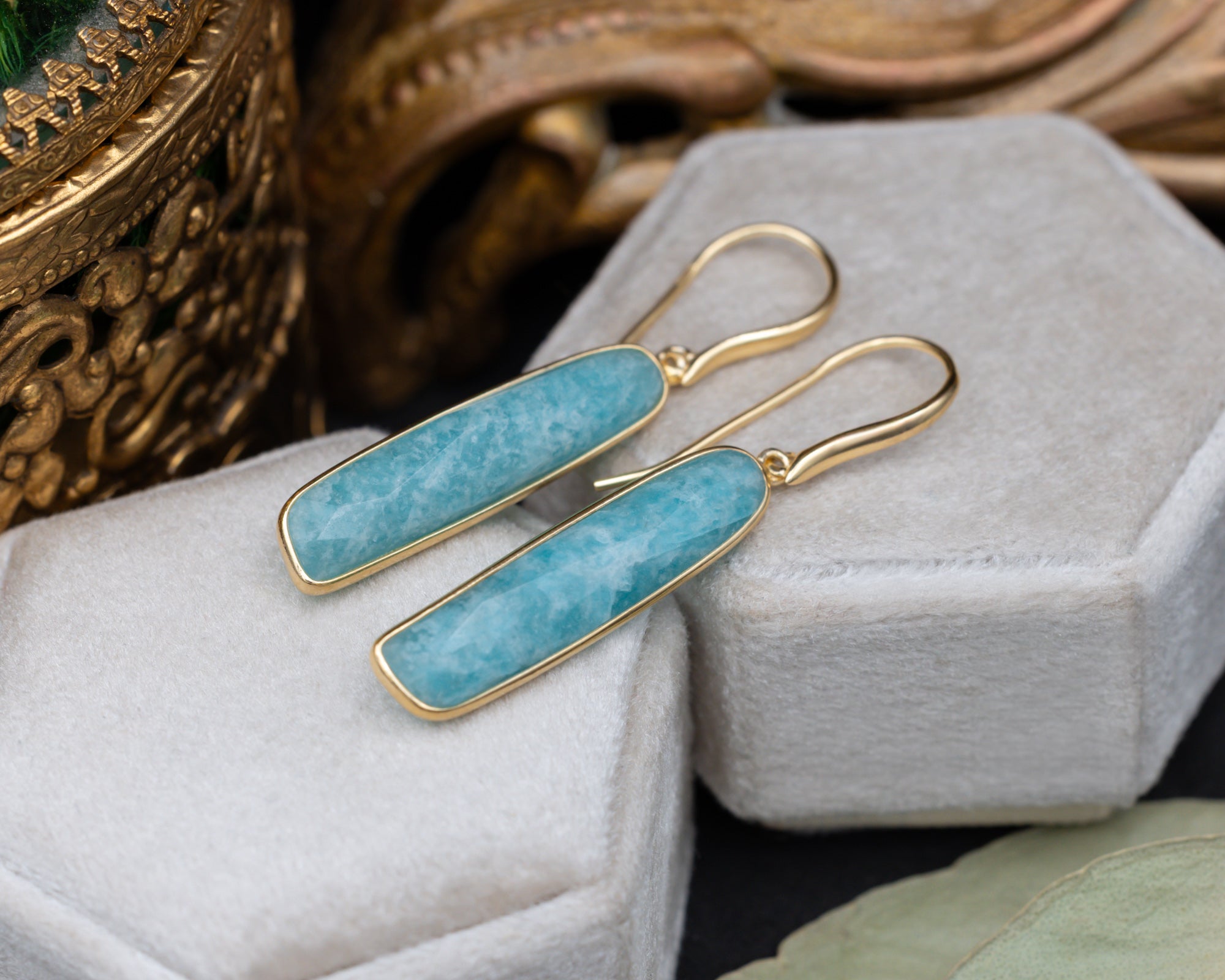 A pair of gold-plated amazonite earrings on a grey cushion with decorative elements in the background