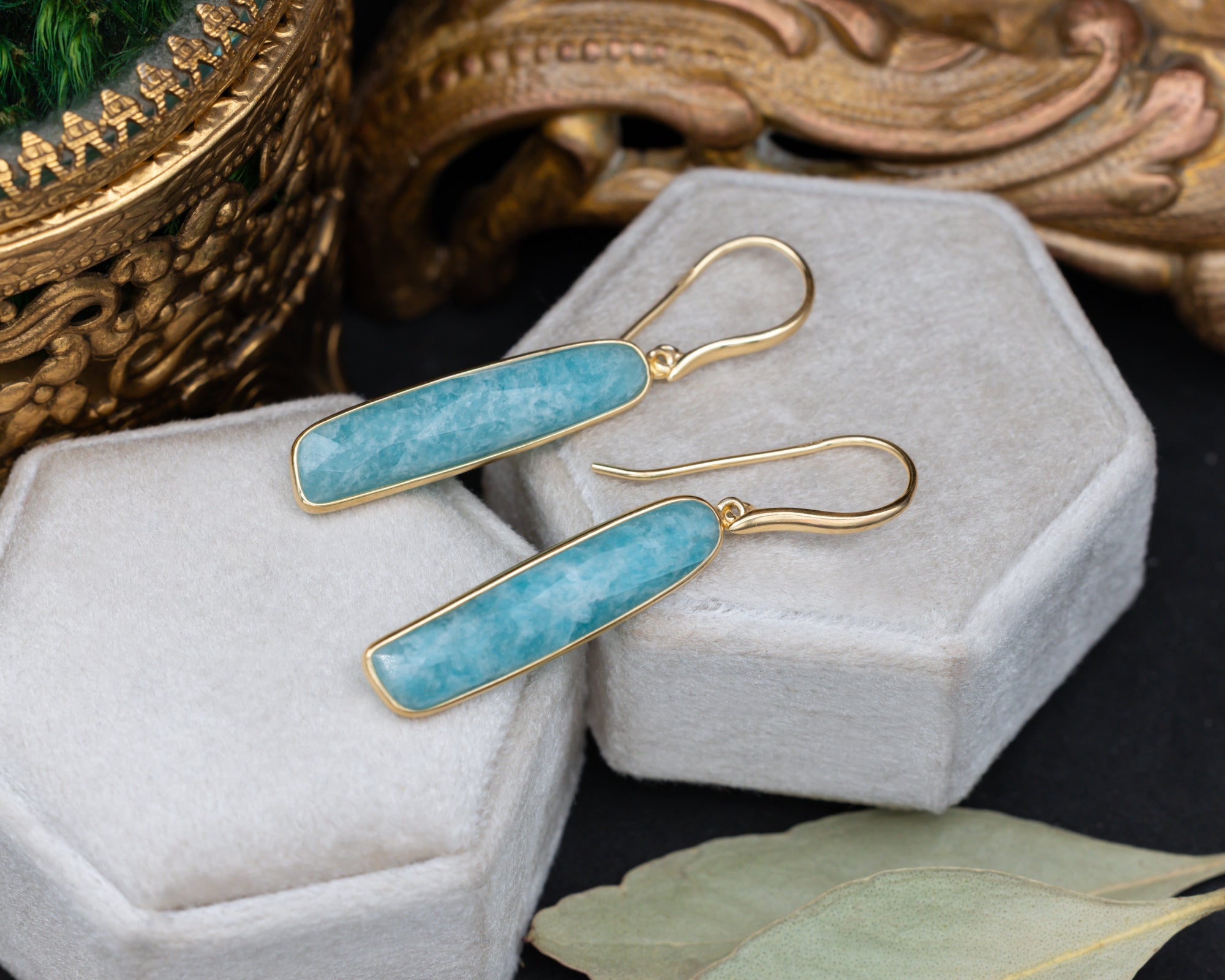 A pair of gold-plated amazonite earrings on a grey cushion with decorative elements in the background
