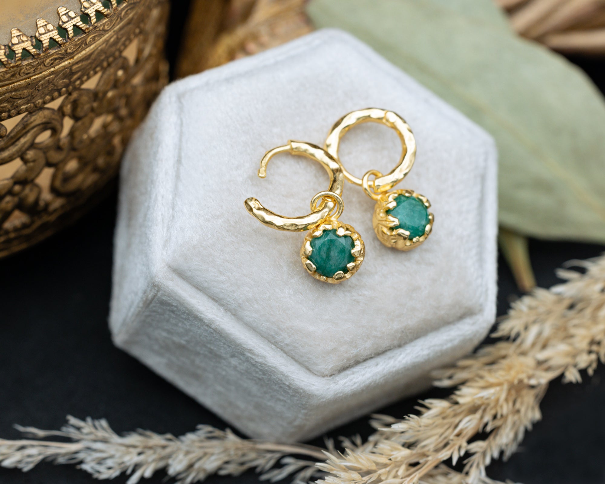 Gold earrings with green emerald on a white hexagonal stand with a natural background