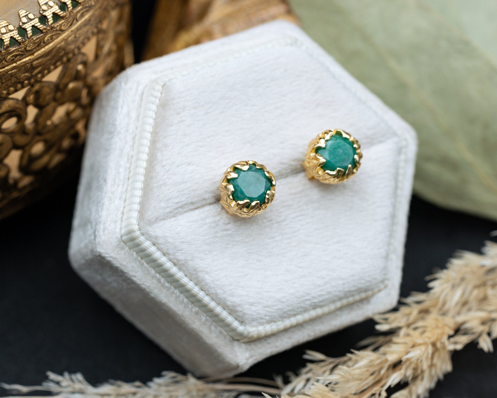 Gold earrings with green emeralds on a white hexagonal stand.