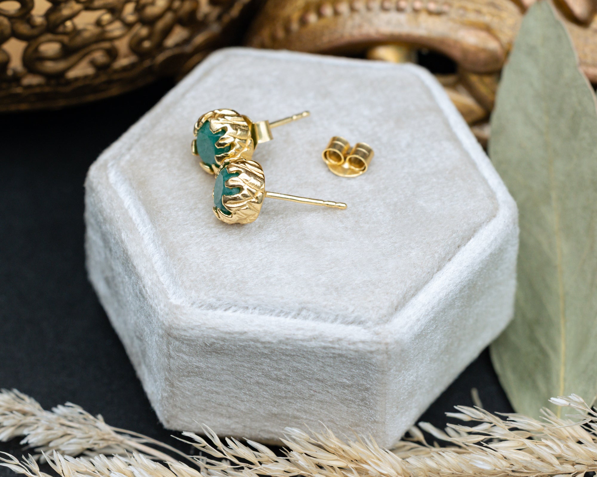 Gold earrings with green emeralds on a hexagonal white velvet box.