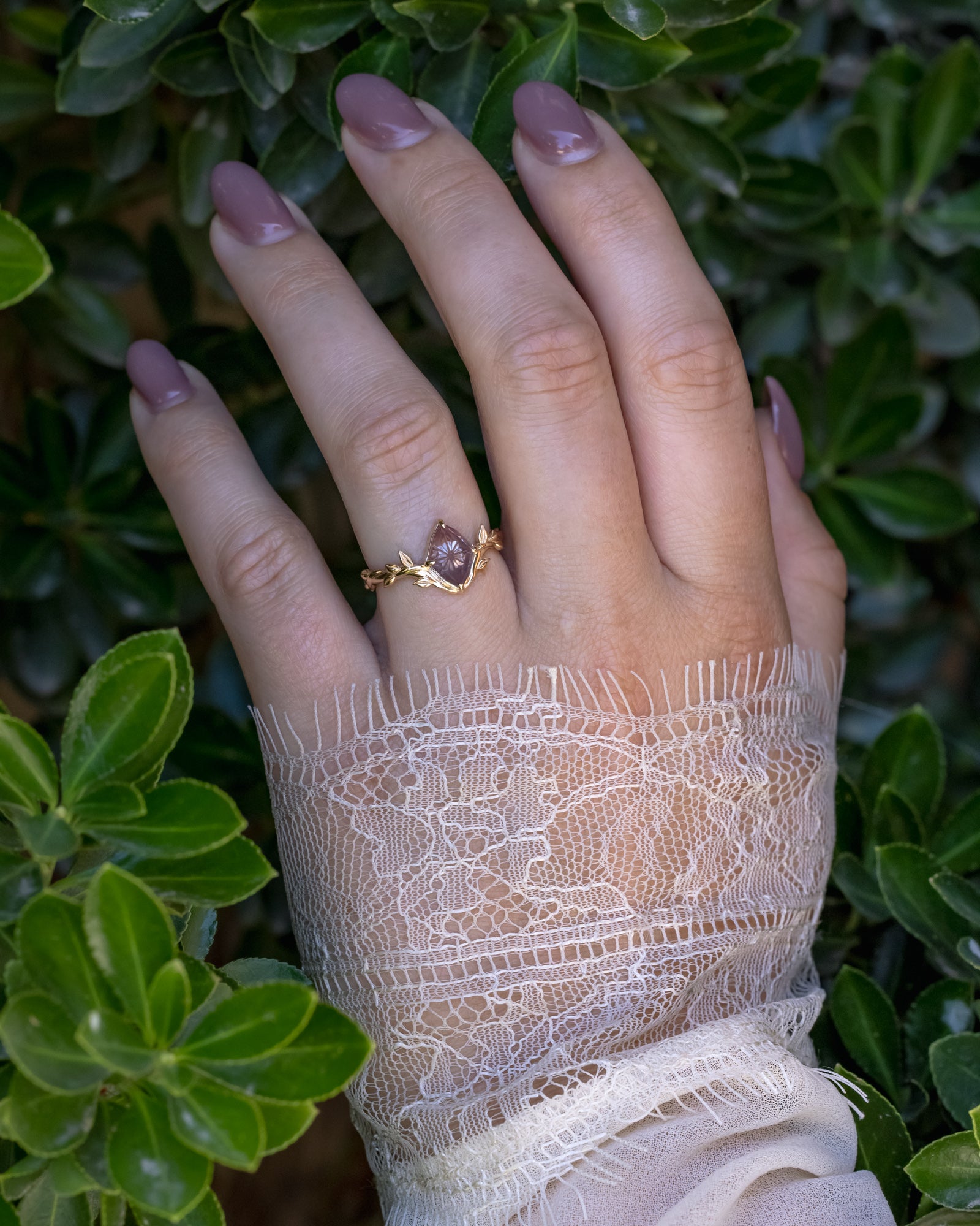 Hand wearing a gold ring with a pink garnet against a green leafy background