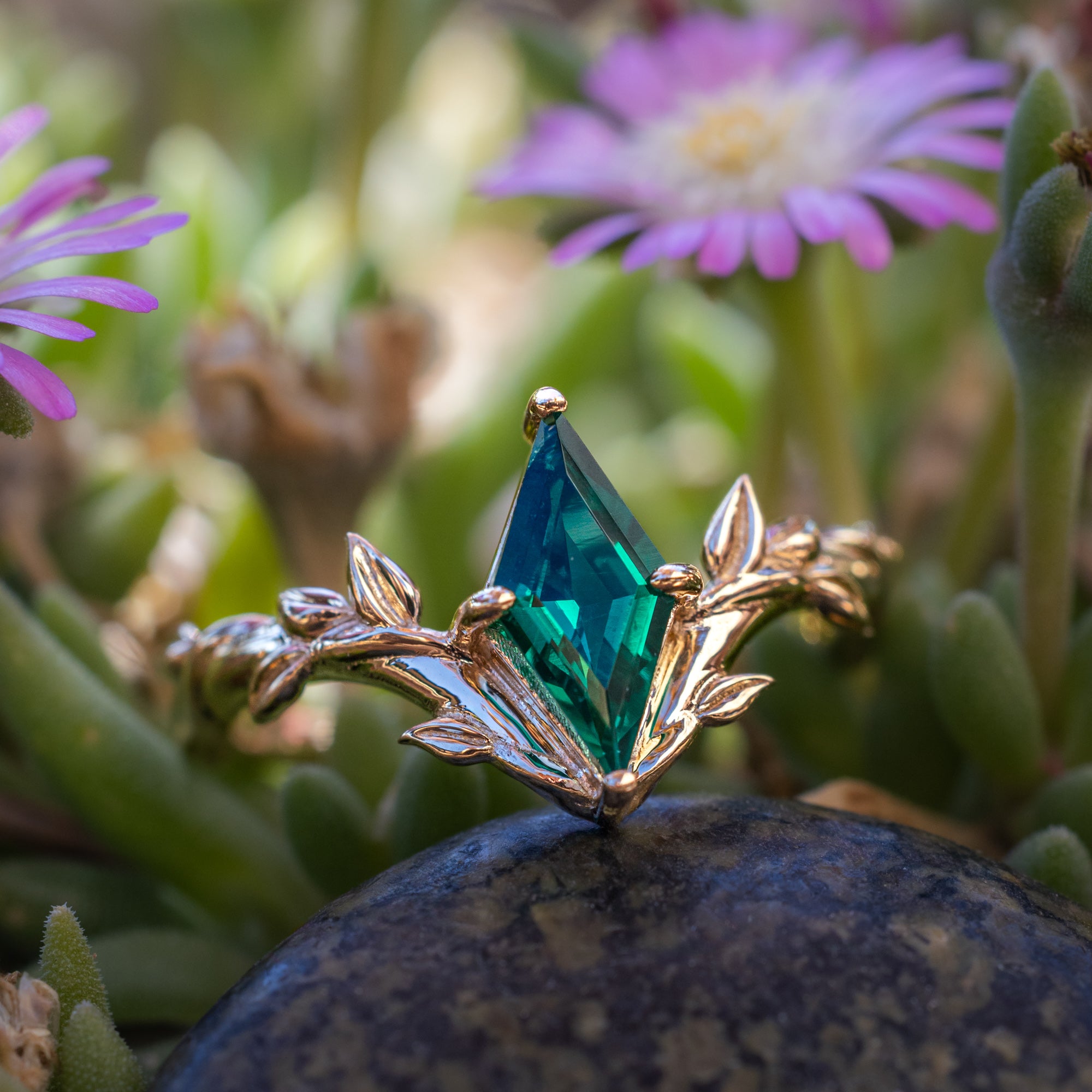 Gold ring with a emerald gemstone on a rock with pink flowers in the background