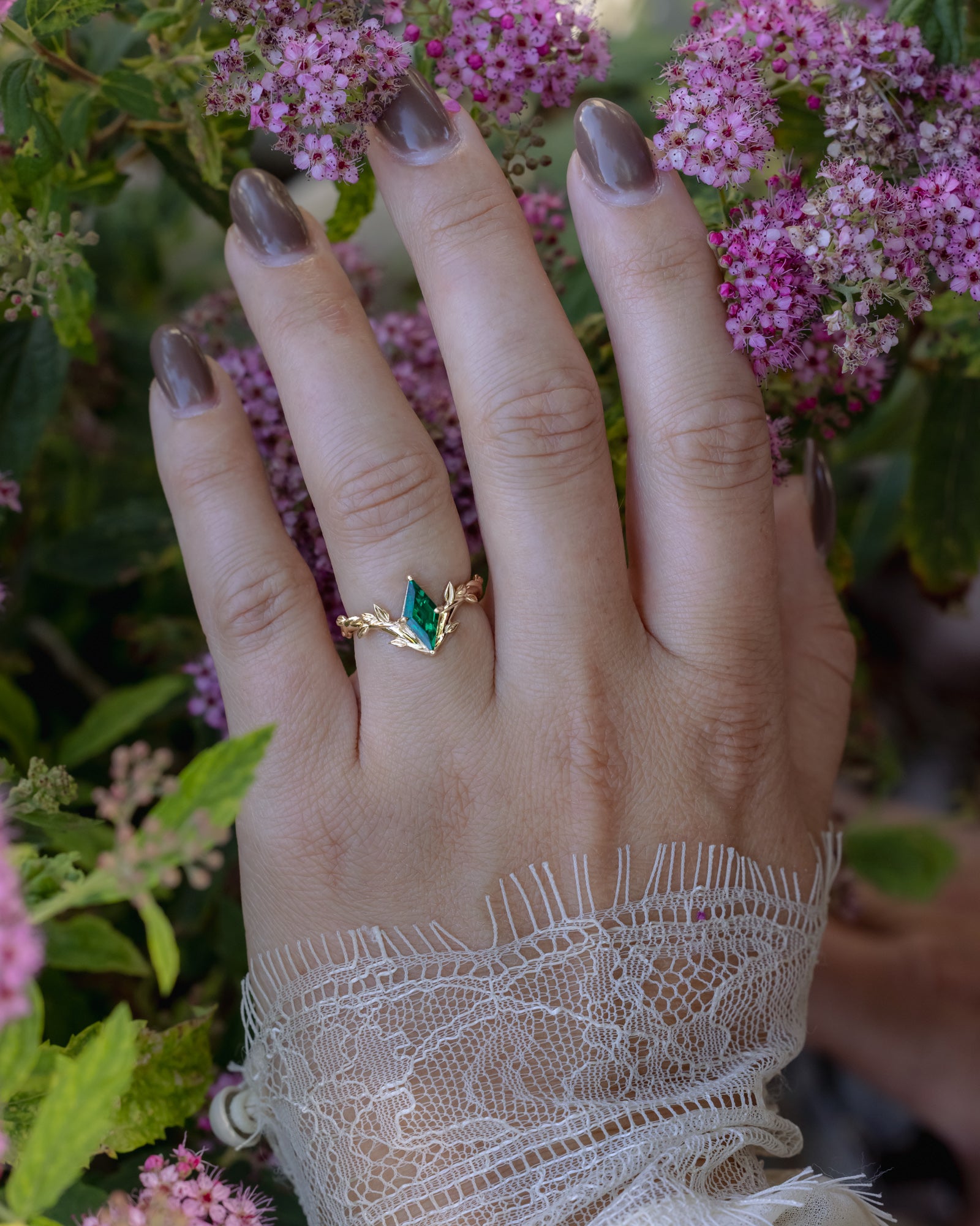 Hand wearing a gold ring with an emerald  center stone against a floral background