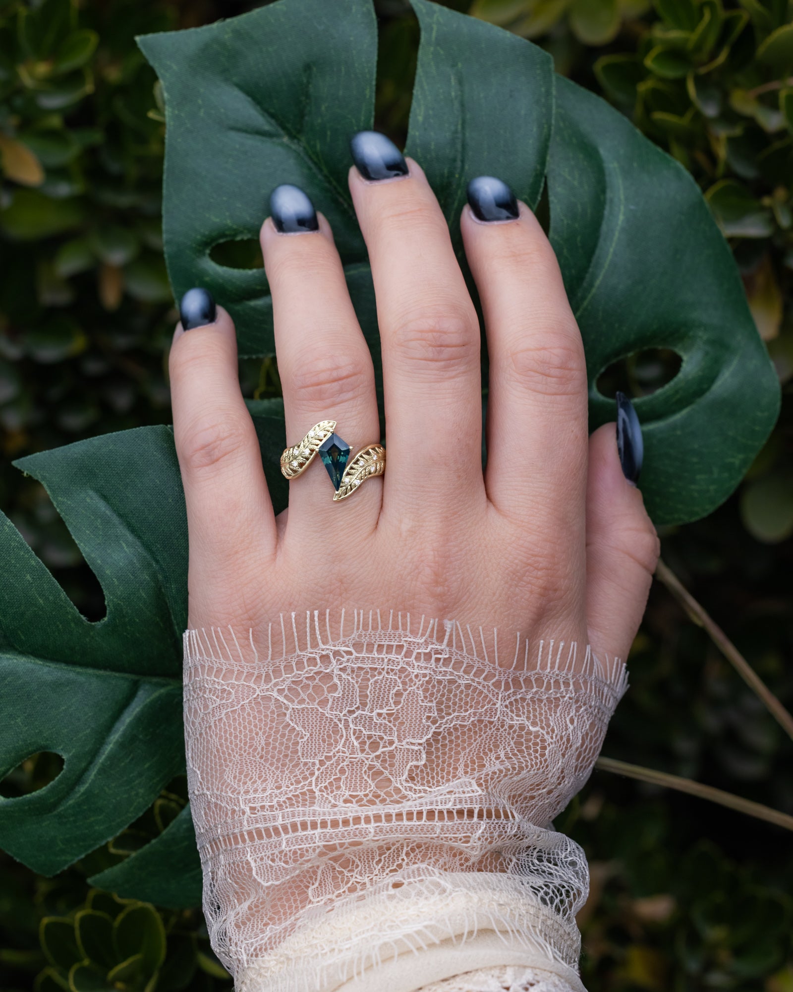Hand wearing a gold ring with a green gemstone against a leafy background