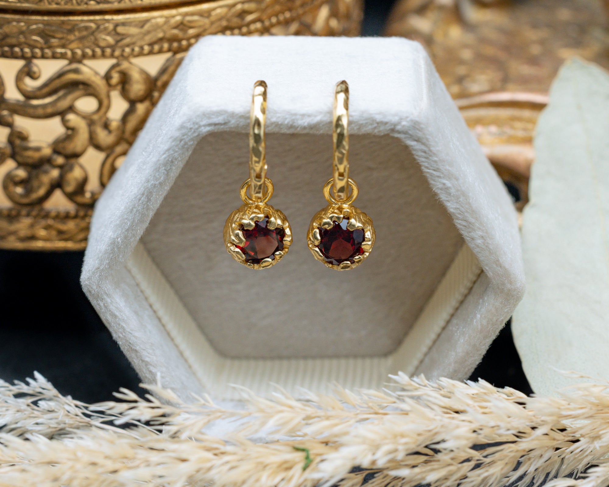 Gold earrings with red garnets in a white hexagonal display box.