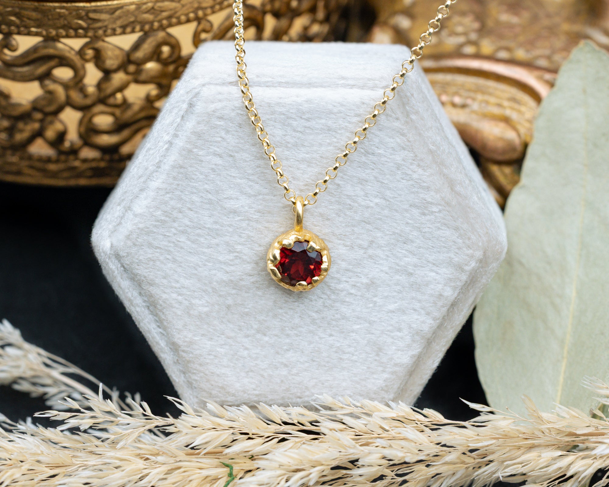 Gold necklace with a red garnet pendant on a white hexagonal stand, surrounded by decorative elements.