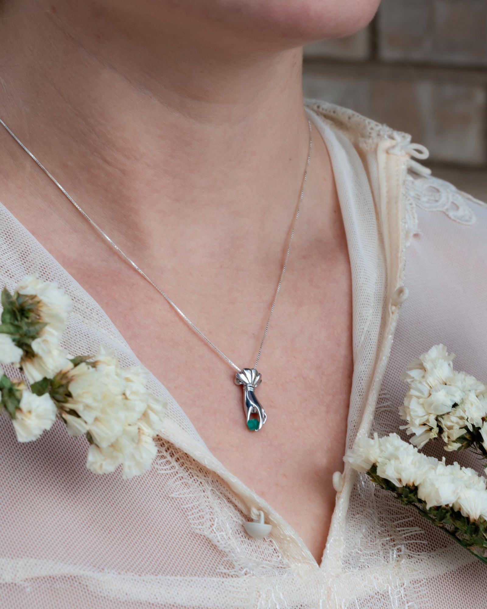 Necklace with a hand pendant worn by a person in a white lace garment, surrounded by white flowers.