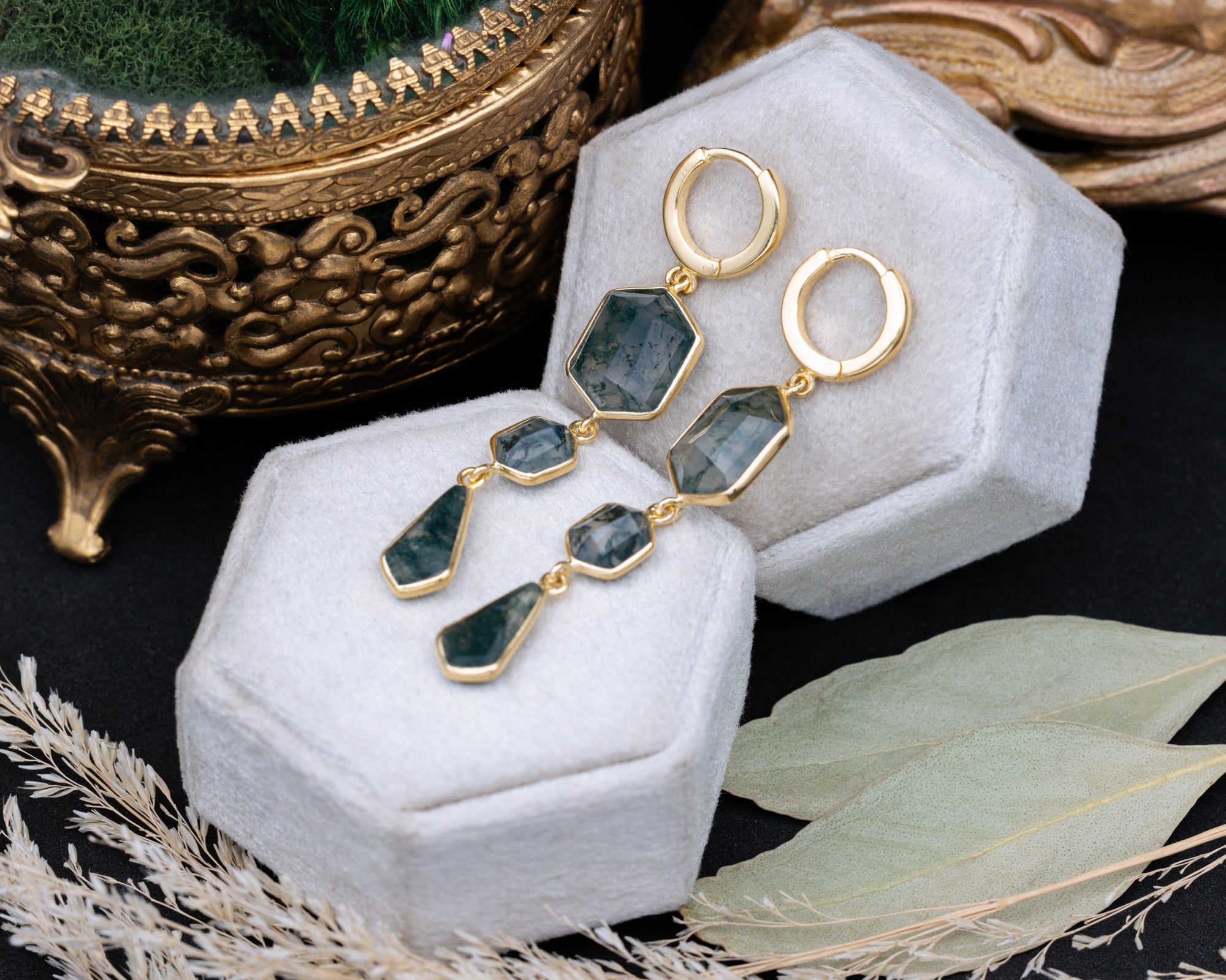 Gold moss agate earrings on a white jewelry box with decorative elements.