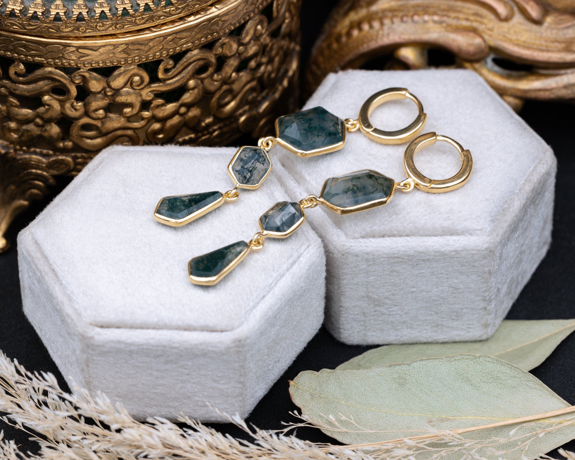 Gold moss agate earrings on a white jewelry box with decorative elements.
