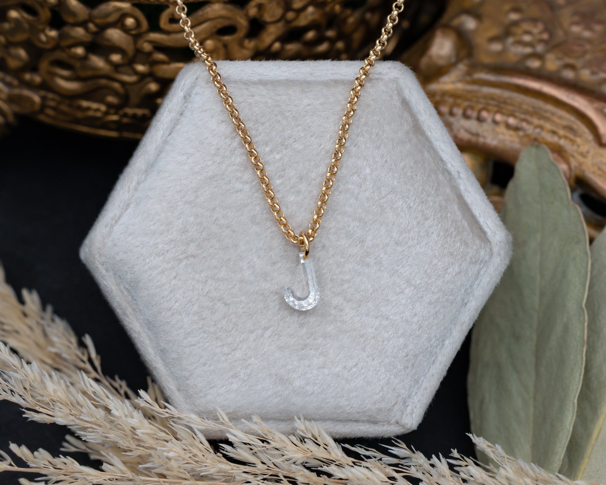 Gold necklace with a 'J' pendant on a hexagonal gray stand.