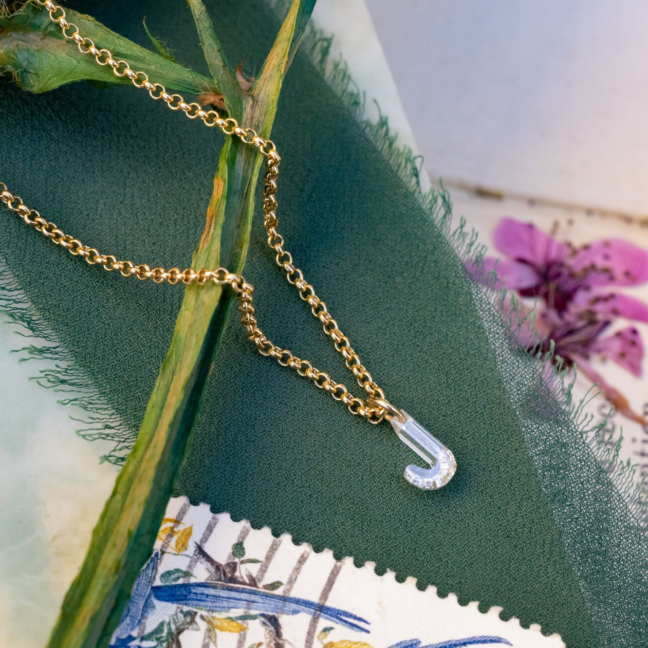 Gold chain with a lab diamond "J" on a green fabric background with floral and text elements.