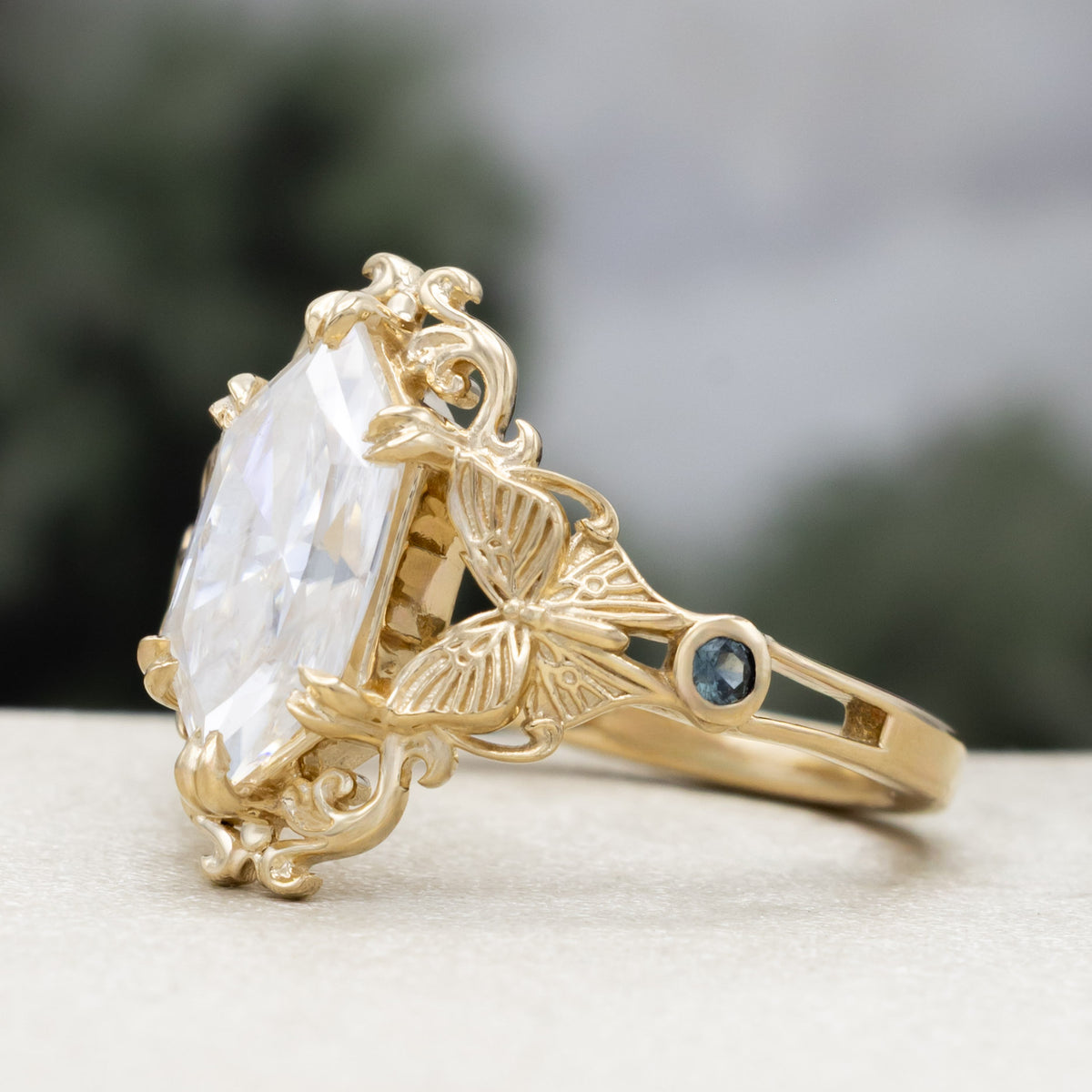Gold ring with moissanite center stone and moth filigree against a blurred green background