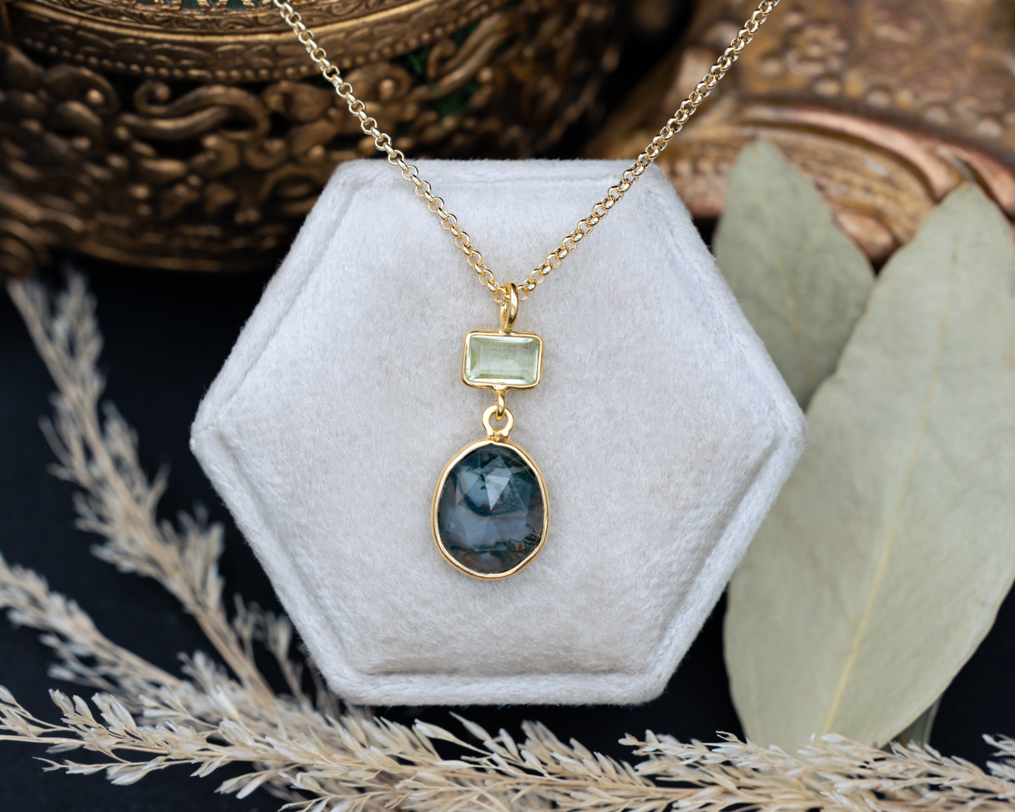 Gold necklace with moss agate & prasiolite pendant on a hexagonal white velvet stand with decorative elements.