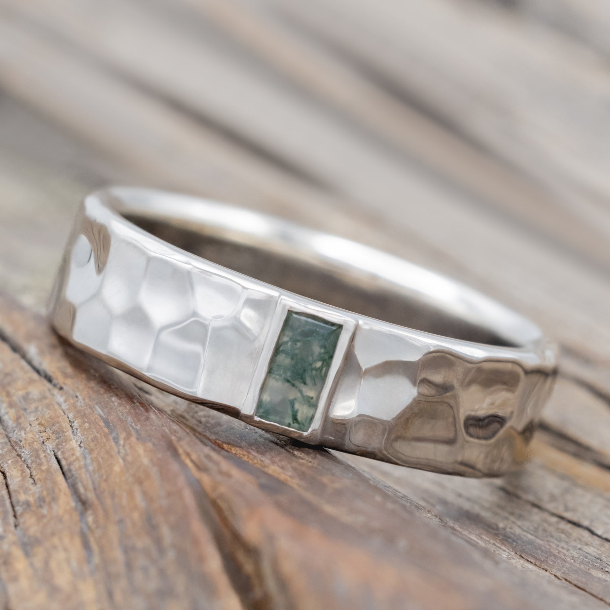 A hammered white gold wedding band with moss agate center stone against a blurred wooden background