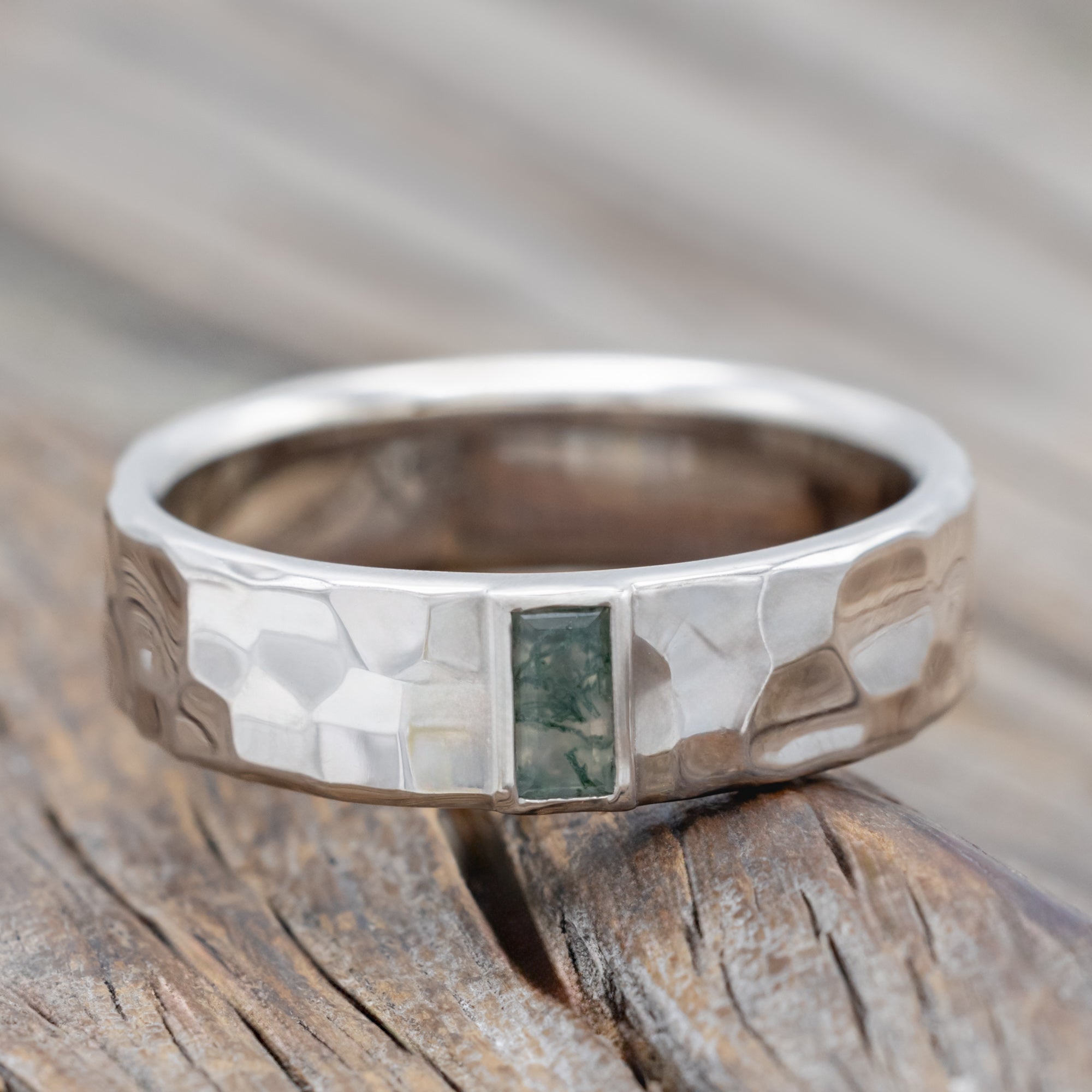 A hammered white gold wedding band with moss agate center stone against a blurred wooden background