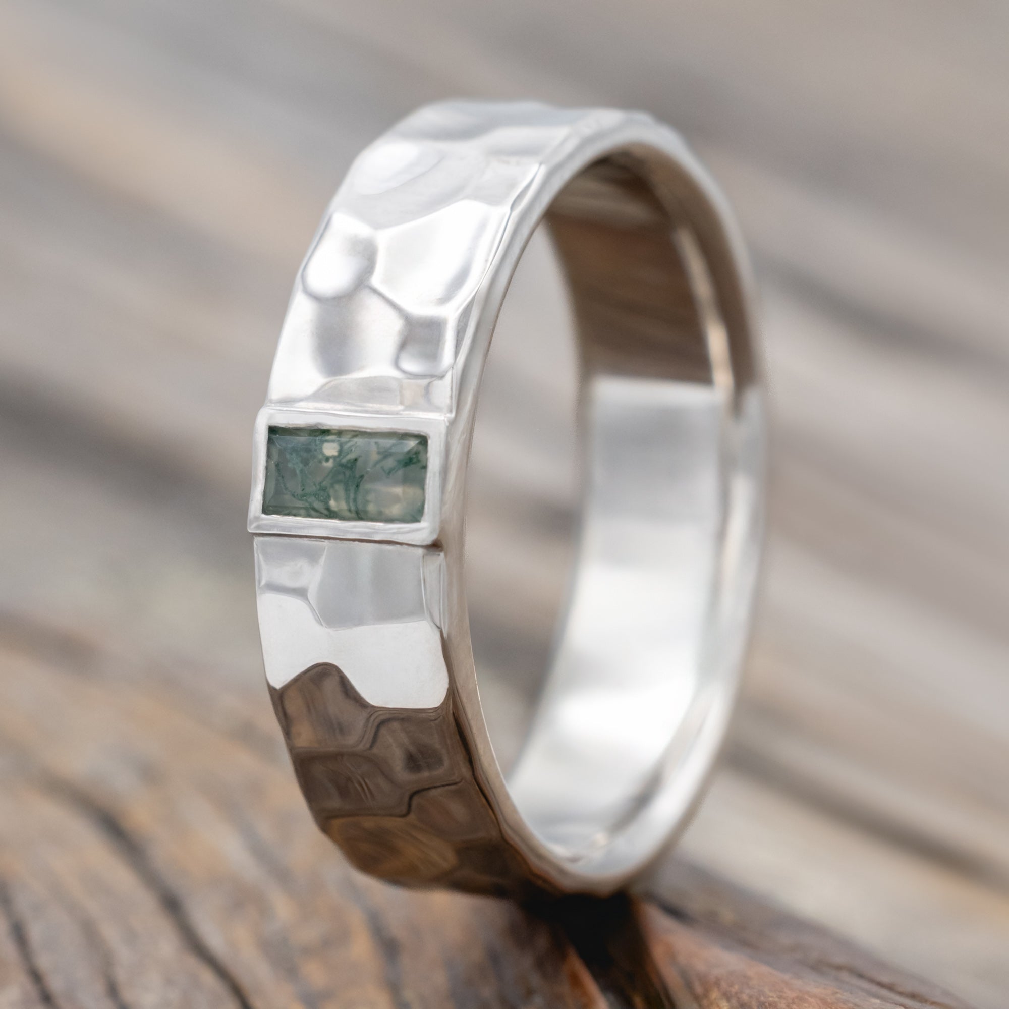 A hammered white gold wedding band with moss agate center stone against a blurred wooden background