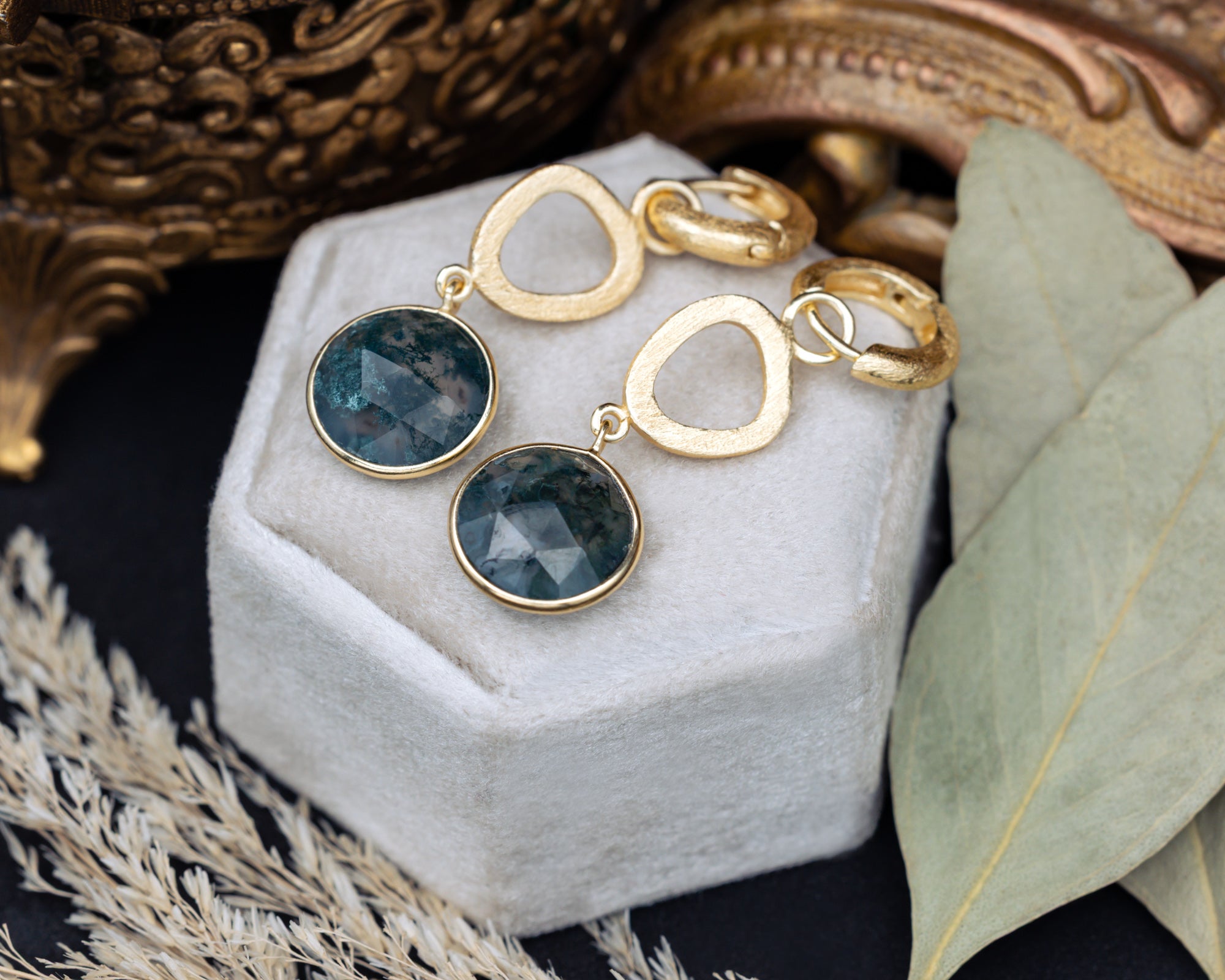 Gold earrings with moss agate gemstones on a white jewelry box with decorative background