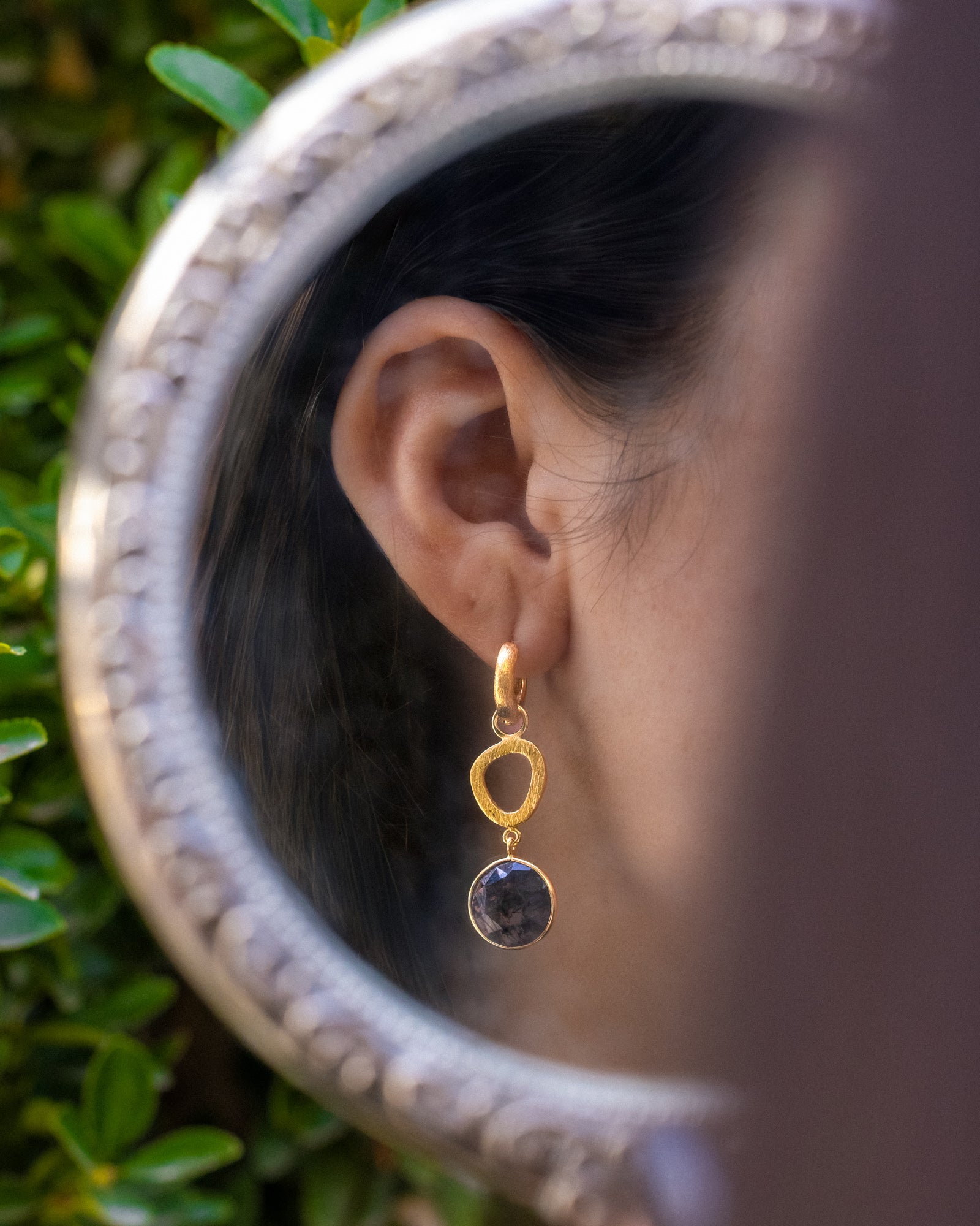 Gold earring with a moss agate worn by a person, with a blurred background.