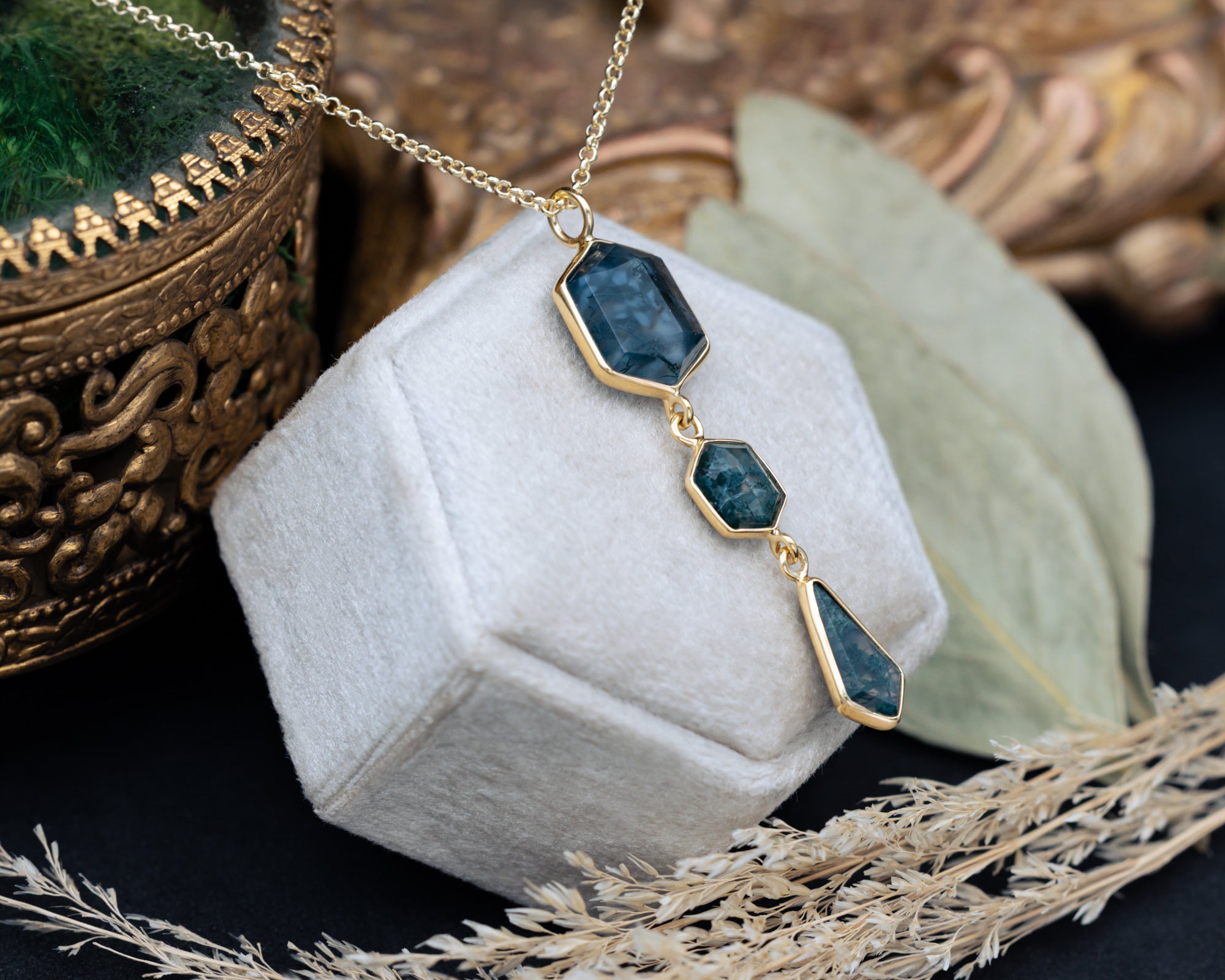 Necklace with moss agate and gold chain on a hexagon cushion with decorative elements in the background