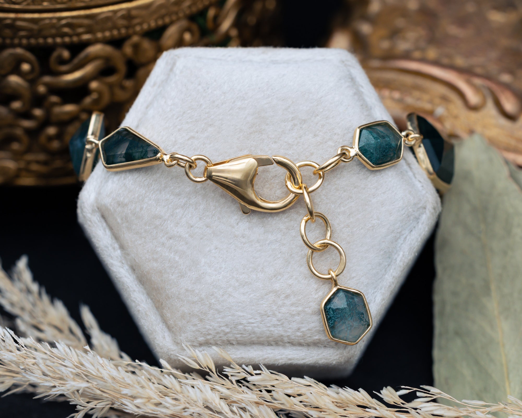 Gold bracelet with moss agate gemstones on a white hexagonal stand with ornate background