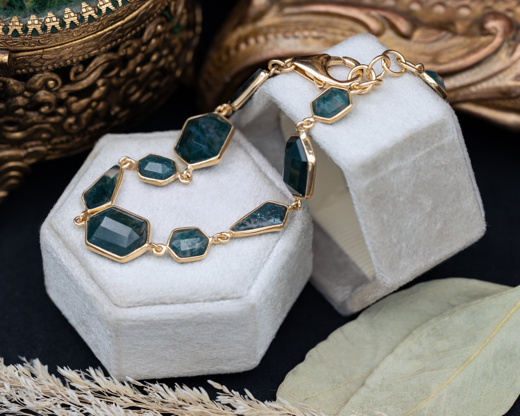 Gold bracelet with moss agate gemstones on a white hexagonal stand with ornate background
