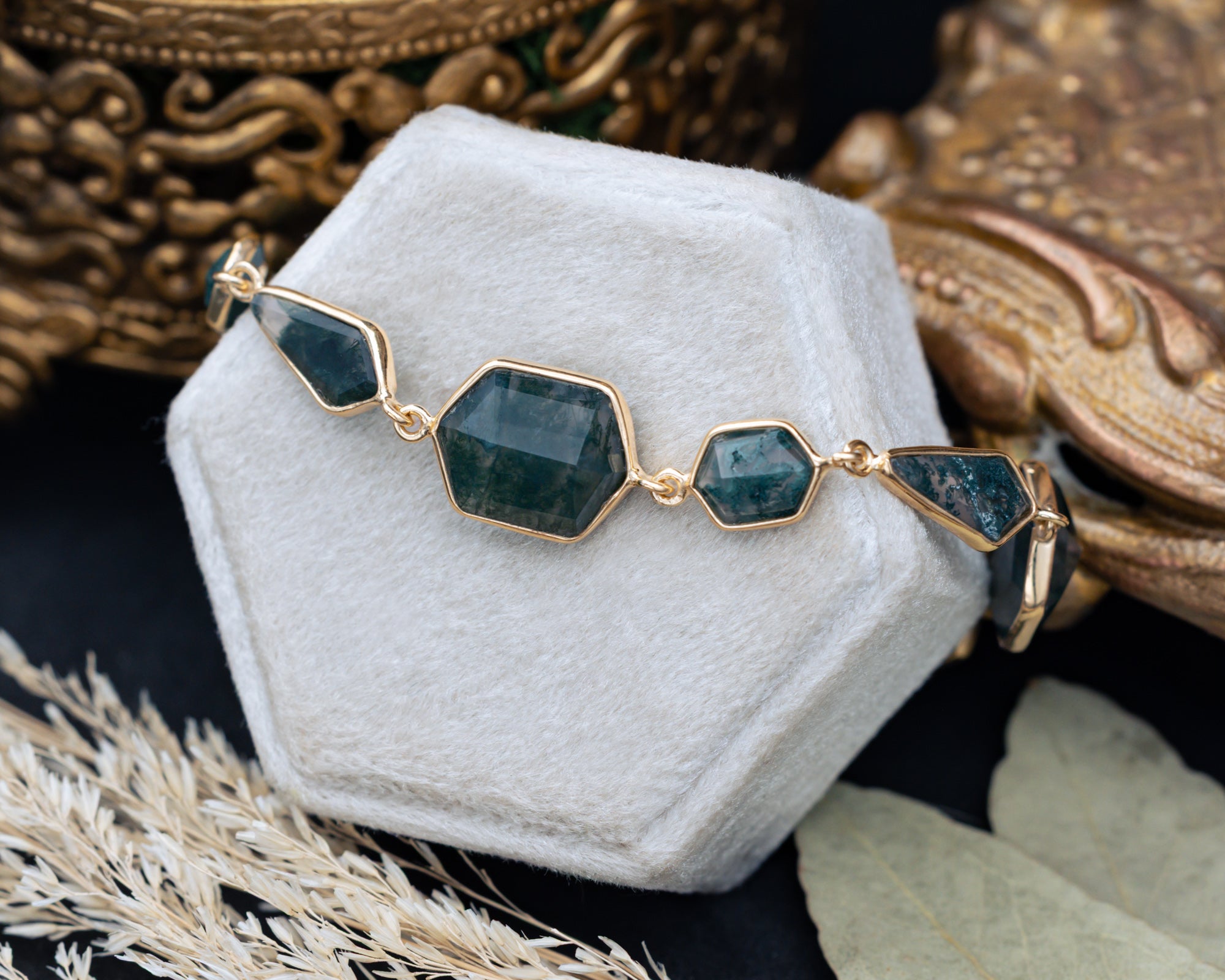 Gold bracelet with moss agate gemstones on a white hexagonal stand with ornate background
