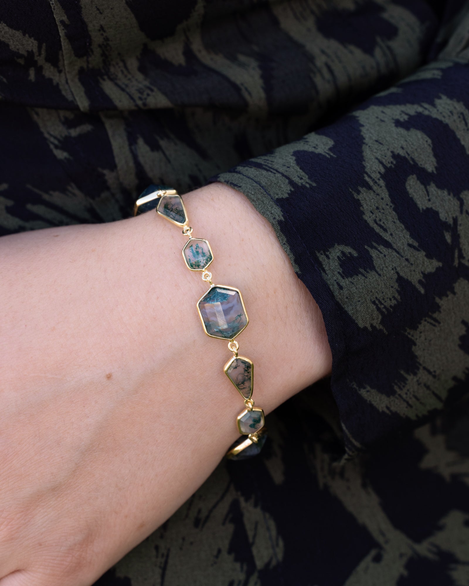 Gold bracelet with gemstone accents on a wrist against a dark patterned fabric background