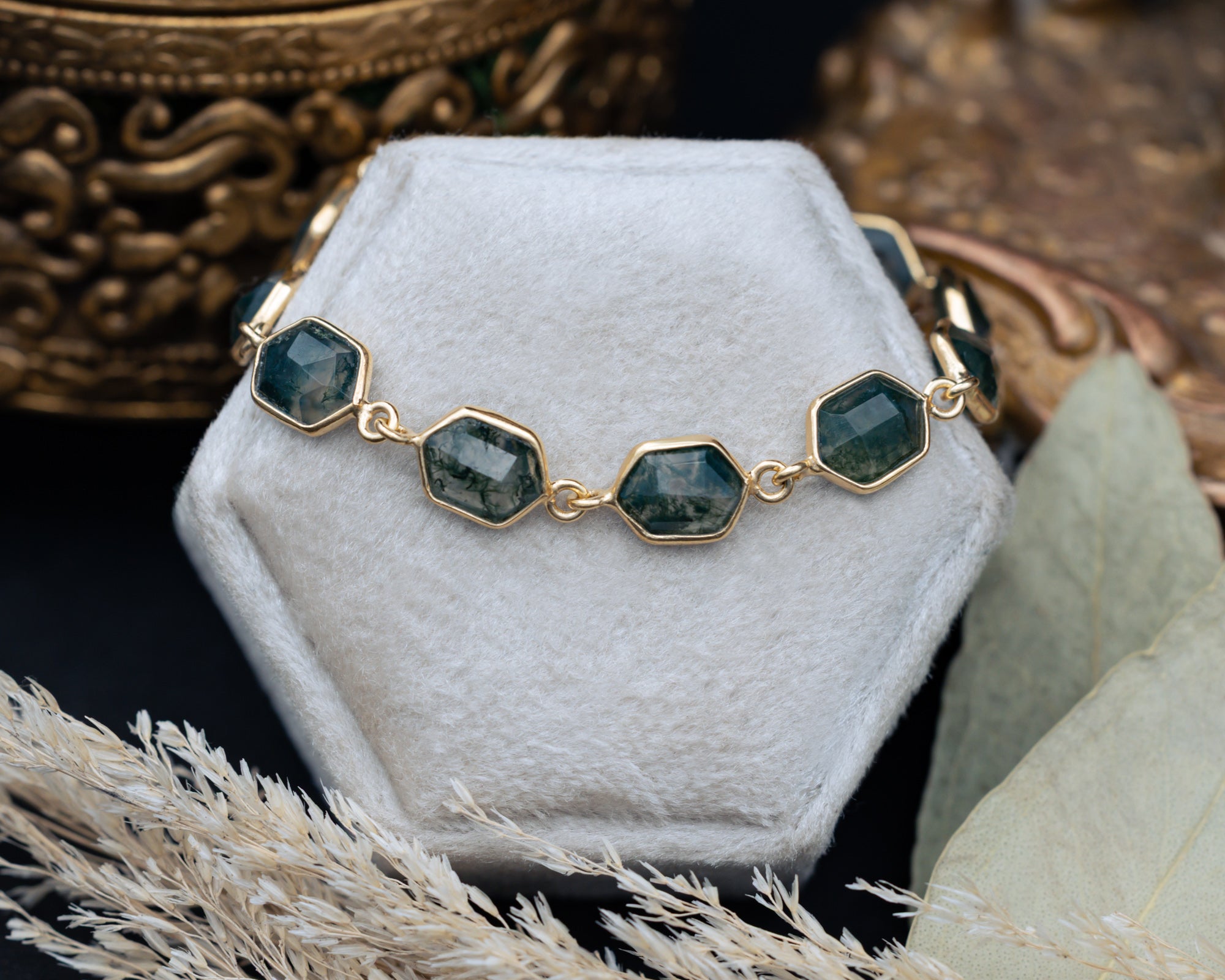 Gold bracelet with hexagon moss agate on a white stand with decorative background