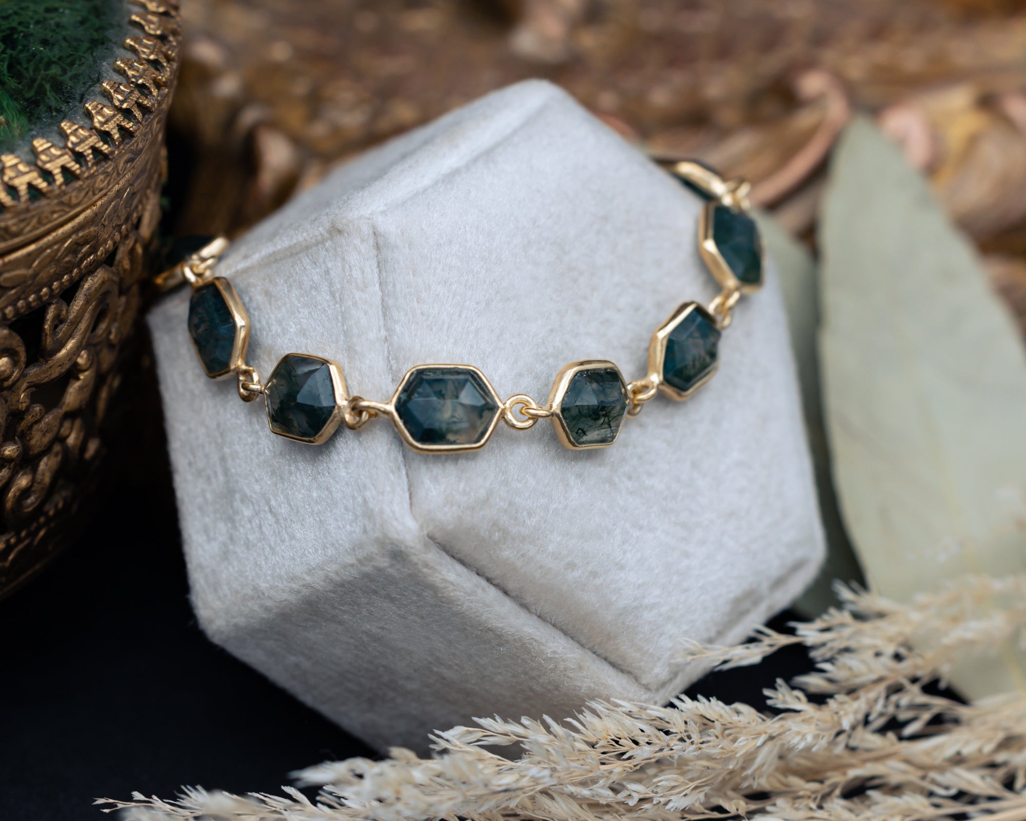Gold bracelet with hexagon moss agate on a white stand with decorative background