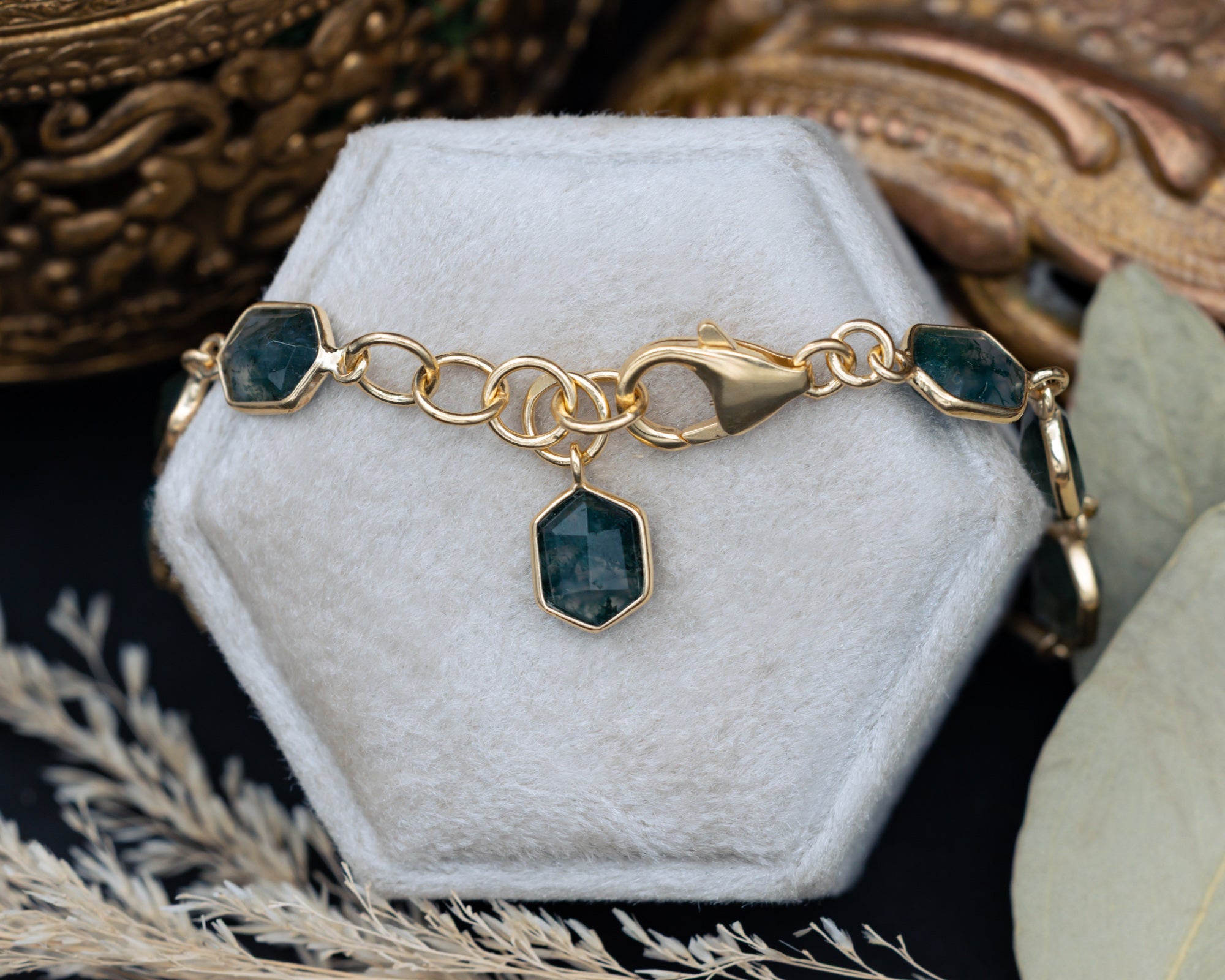 Gold bracelet with hexagon moss agate on a white stand with decorative background