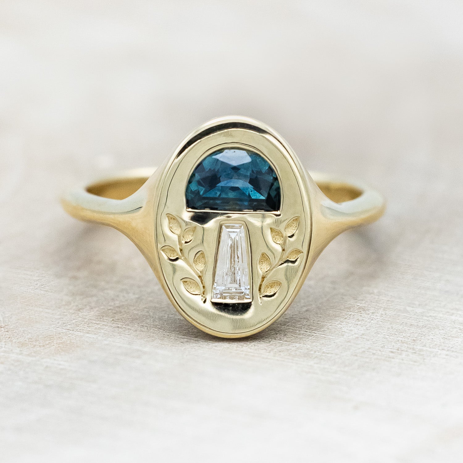 A 14K yellow gold signet ring with a half moon gem cap, tapered baguette diamond stem, and vines engraved on both sides.