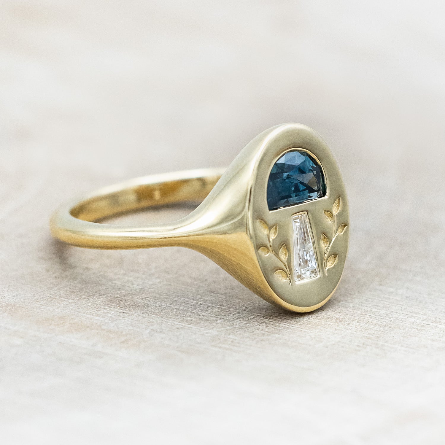 SIde profile of a 14K yellow gold signet ring with a half moon gem cap, tapered baguette diamond stem, and vines engraved on both sides.