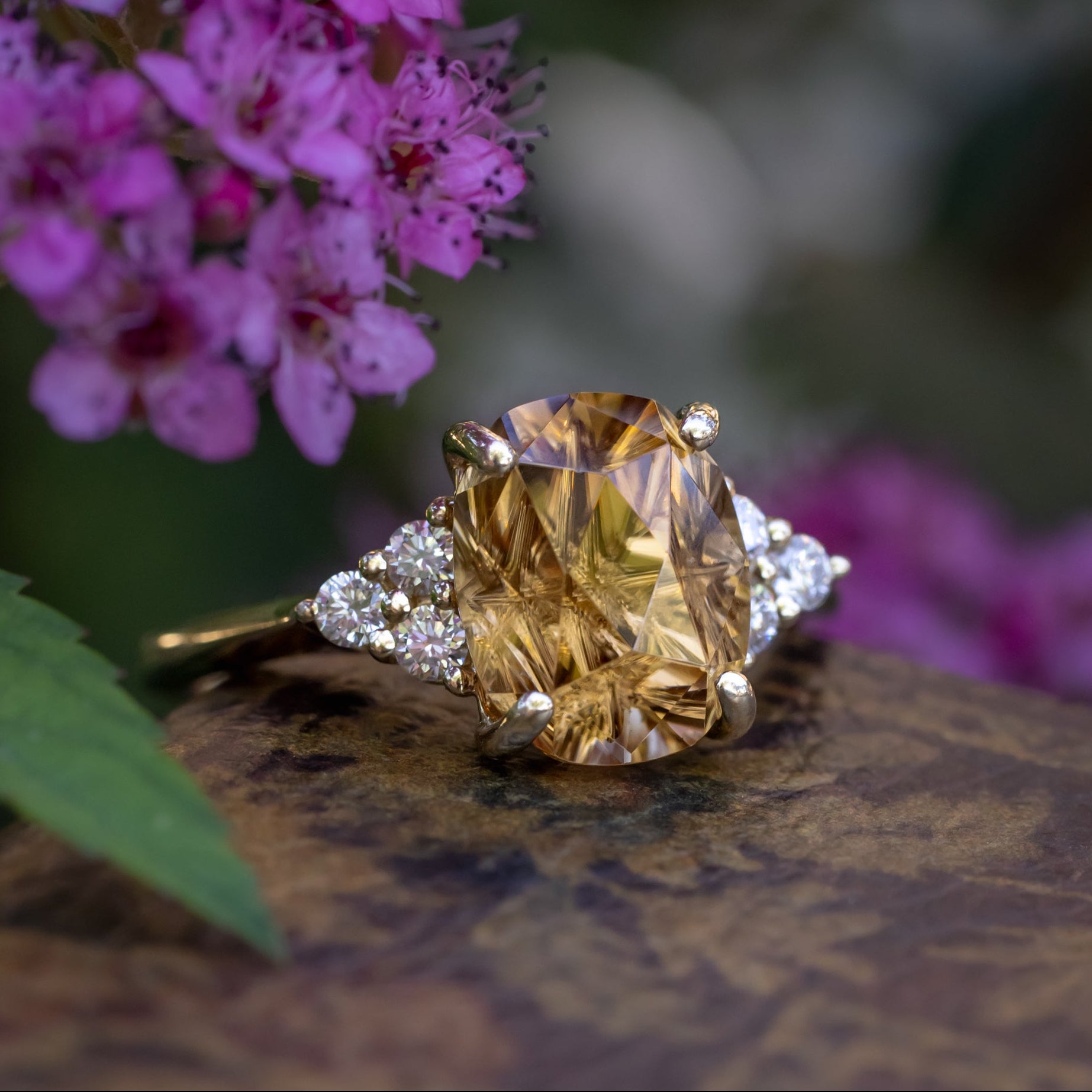 Gold ring with a yellow zircon and diamond accents, placed on a textured surface with pink flowers in the background.