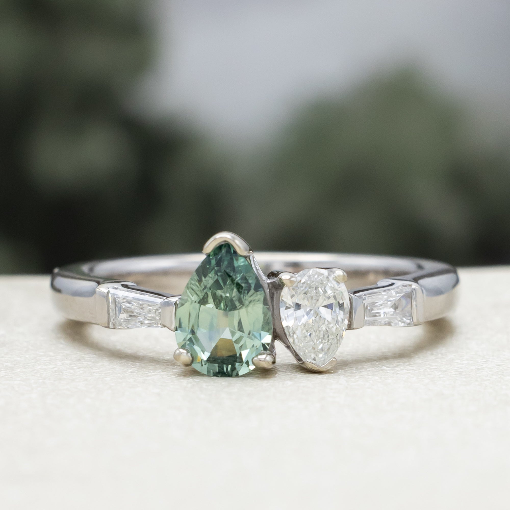 White gold ring with a pear sapphire and diamond next to each other with diamond accents against a blurred background