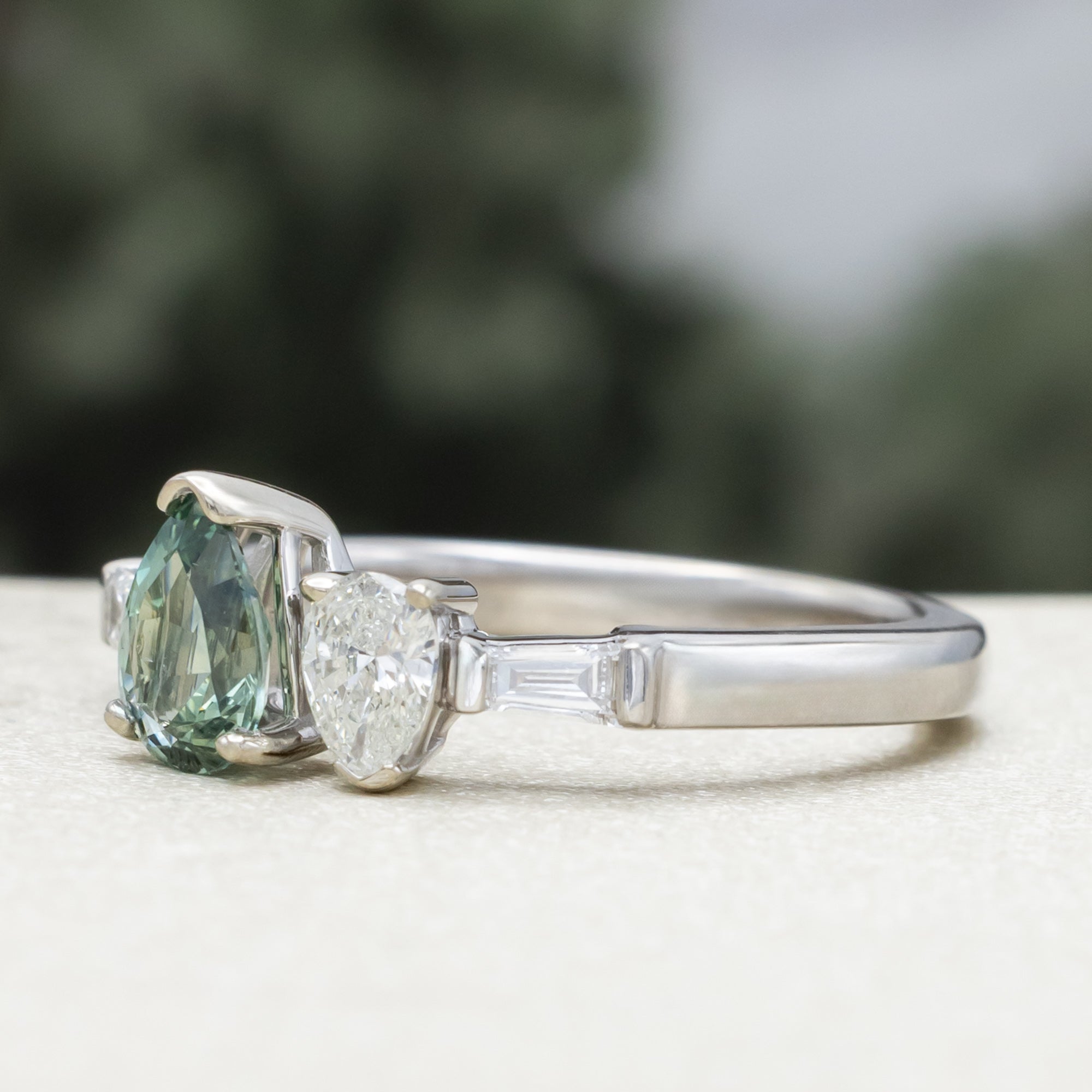 White gold ring with a pear sapphire and diamond next to each other with diamond accents against a blurred background