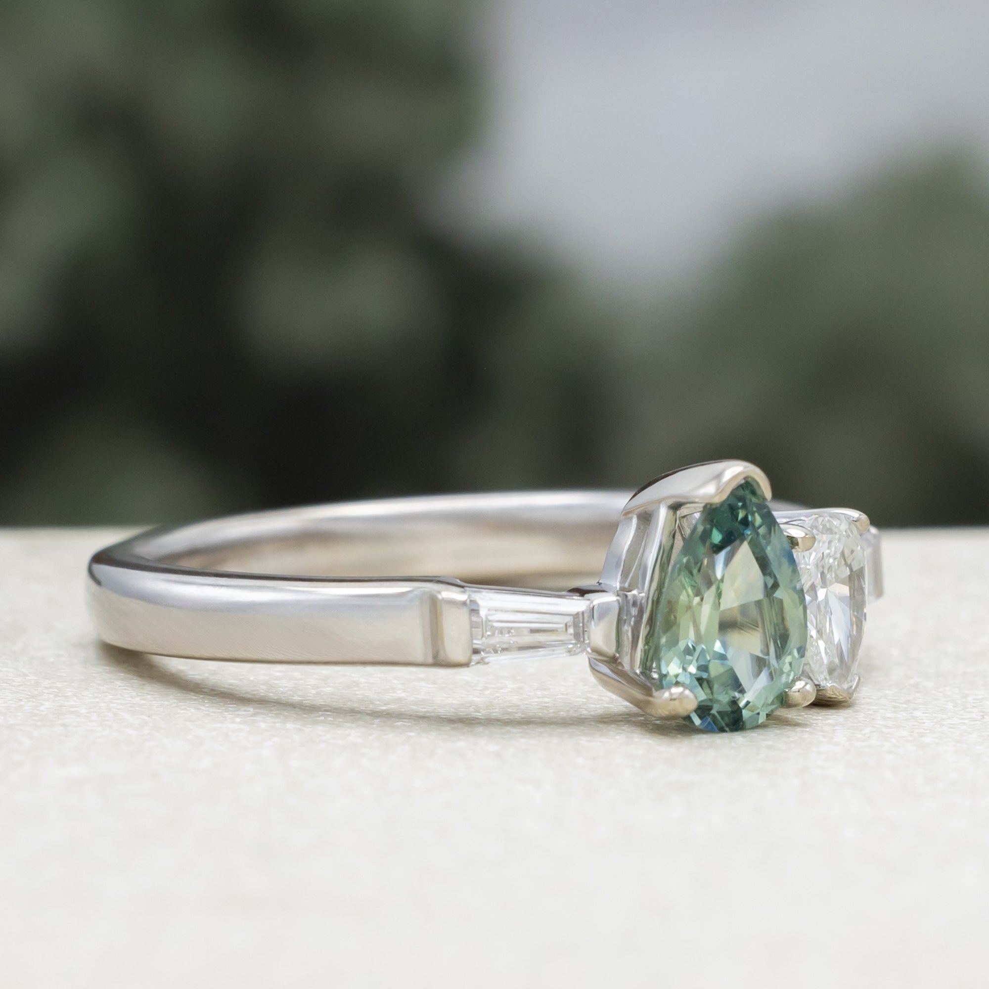 White gold ring with a pear sapphire and diamond next to each other with diamond accents against a blurred background