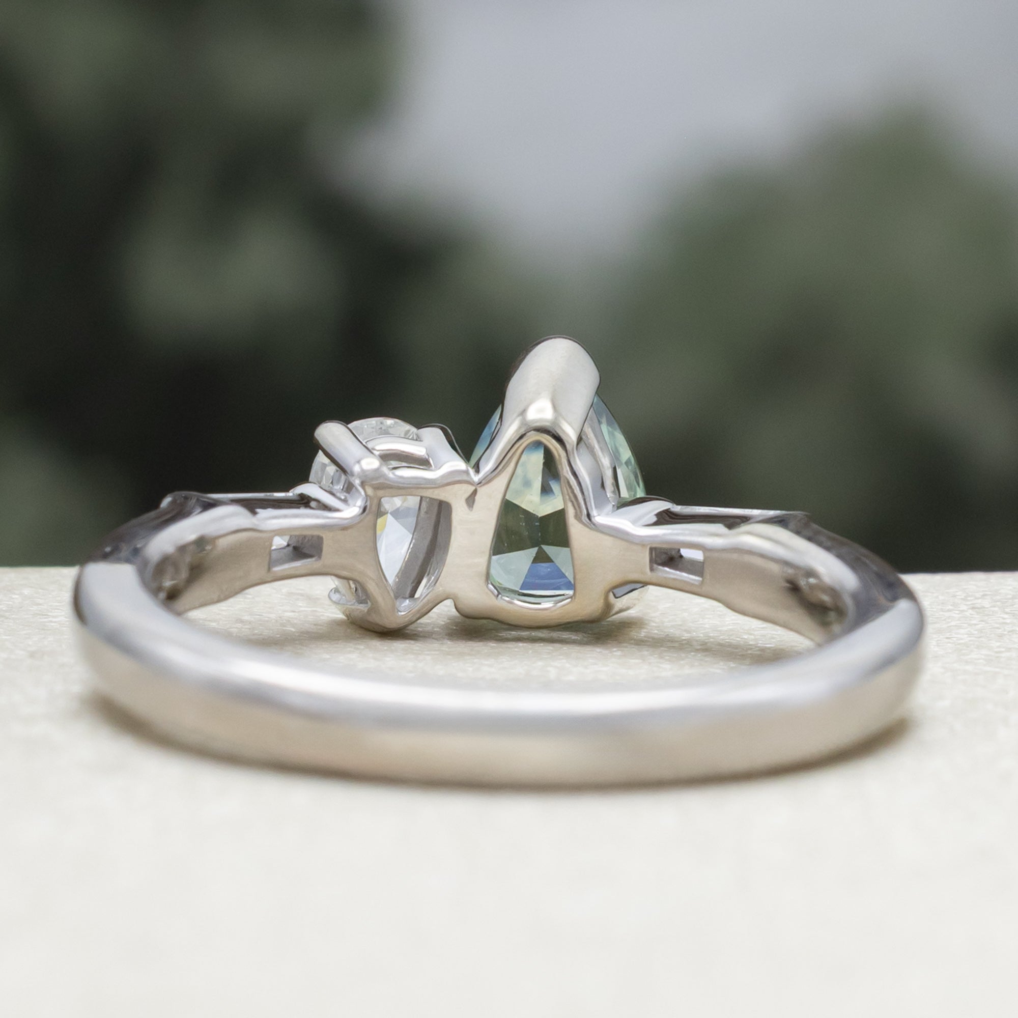 White gold ring with a pear sapphire and diamond next to each other with diamond accents against a blurred background