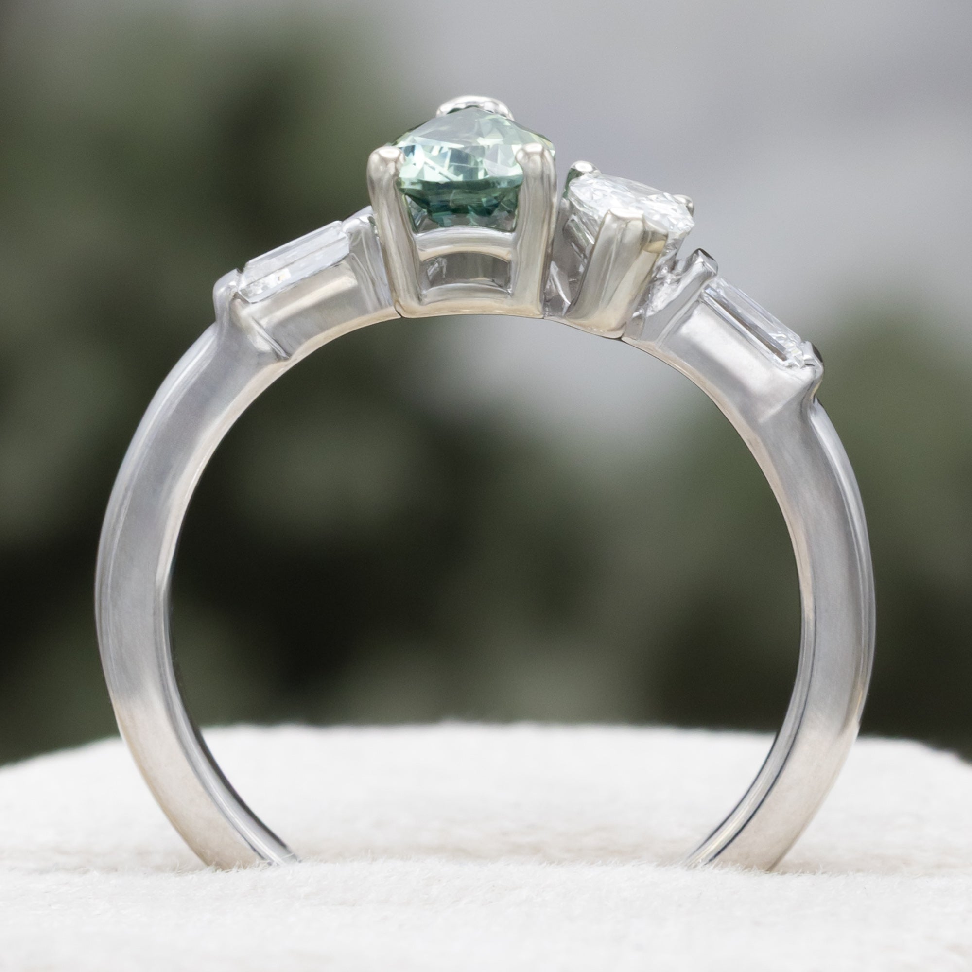 White gold ring with a pear sapphire and diamond next to each other with diamond accents against a blurred background
