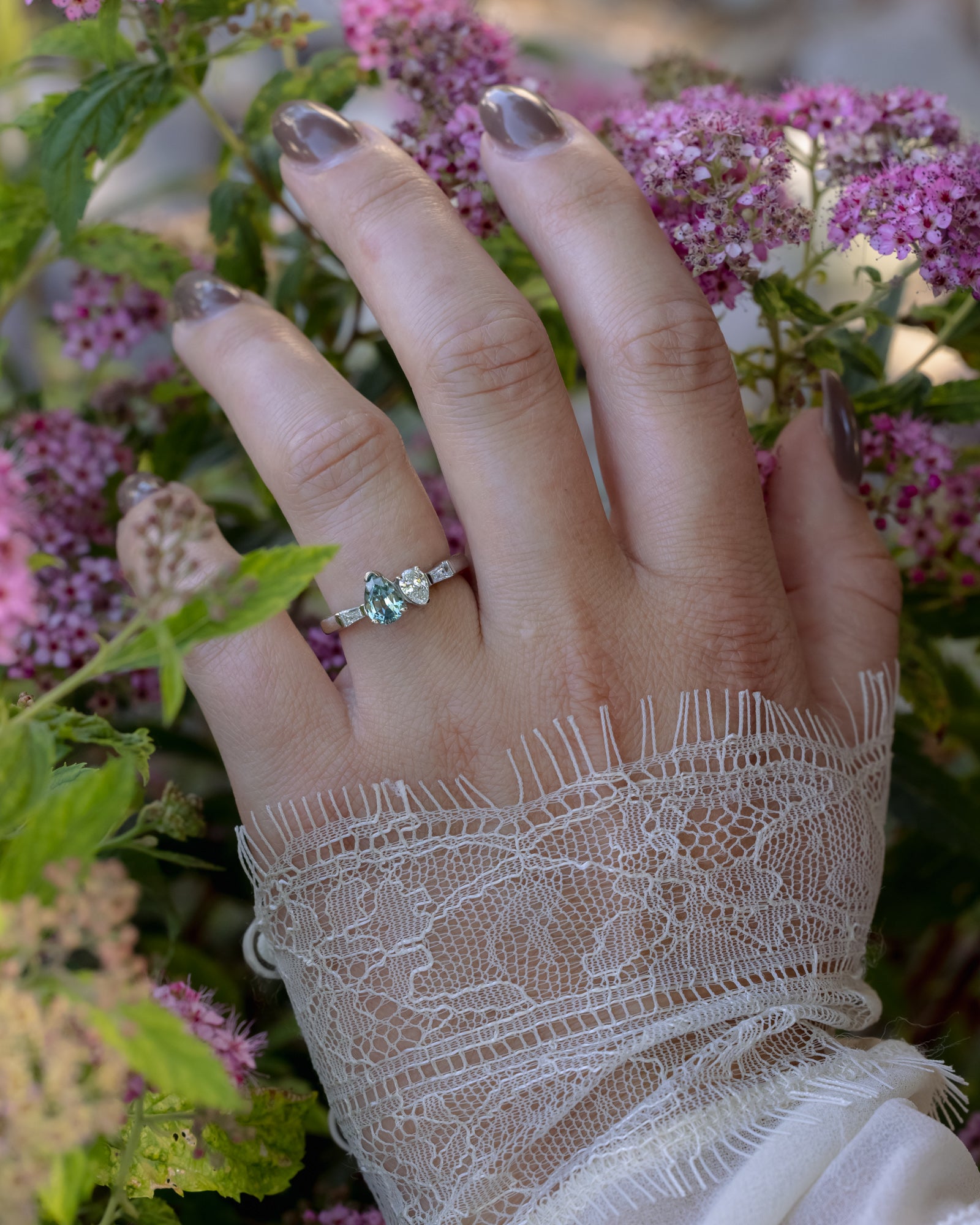 Hand wearing a ring with a Montana sapphire and diamond accents, surrounded by pink flowers